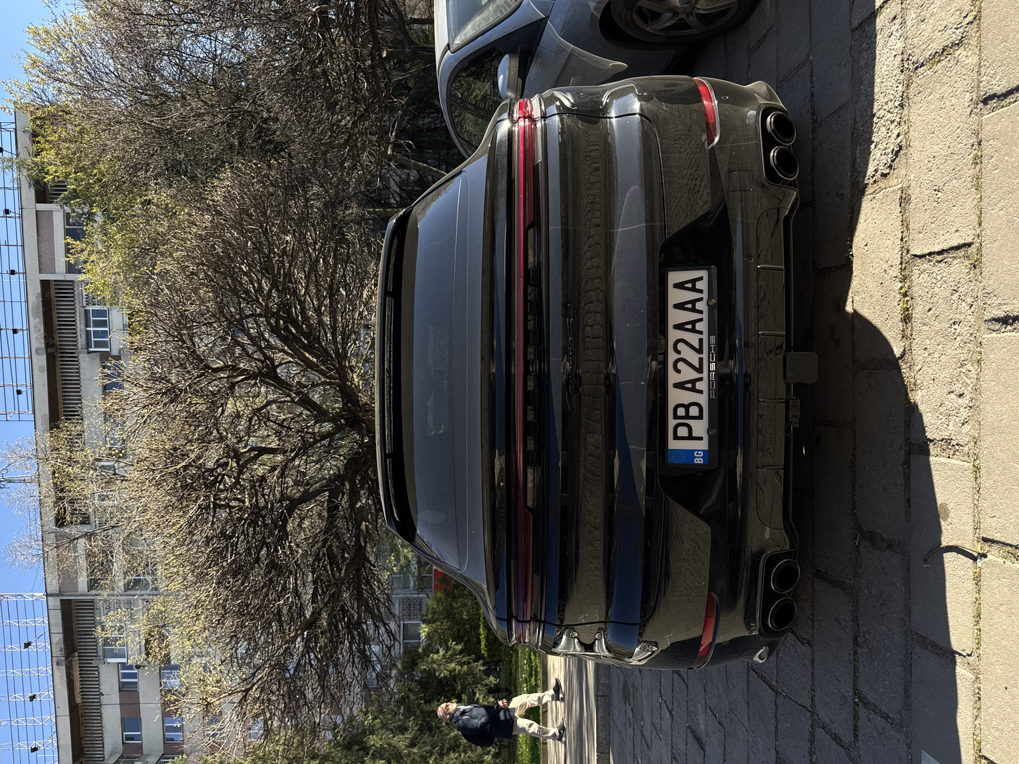 The image shows the rear of a dark-colored Porsche, parked on a cobblestone street in Plovdiv, Bulgaria. The car has a Bulgarian license plate, "PB A22AAA". To the left of the vehicle, a leafless tree is prominent, in front of a multi-story building with rows of windows. A person is walking on the sidewalk near the car, dressed in a dark jacket and light-colored pants. The environment suggests a sunny day in an urban setting.