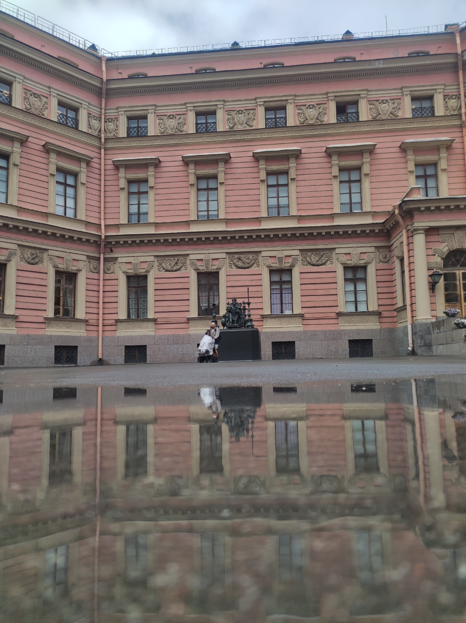 The image depicts a view of the Mikhailovsky Castle (also known as the Engineers' Castle) in Saint Petersburg, Russia, reflected in a puddle on the ground. The castle is a large, pink, classical building with numerous windows and decorative architectural elements. The sky above is overcast, contributing to the muted tones of the scene. In the foreground, a reflection of the castle and the sky is visible on the wet surface of the ground. Two people are present near a statue in front of the castle, with one person appearing to push a stroller.