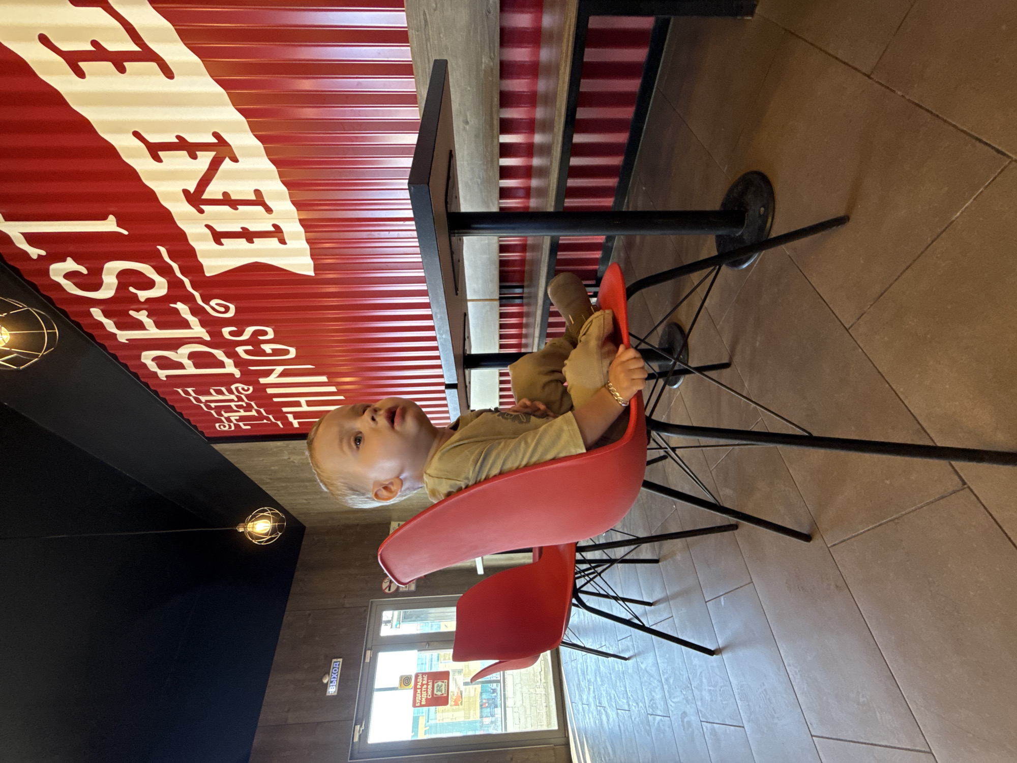 The image captures the interior of a casual dining establishment, likely a fast-food restaurant or cafe, located in Rusanovo, Russia. A young, blonde-haired child, appearing to be a toddler, sits alone in a bright red, mid-century modern-style chair with a black wireframe base. The child wears a khaki-colored long-sleeved shirt and pants, with a thin bracelet on the left wrist, and looks upwards and to the right with a slightly open mouth.
The right wall features prominent red corrugated metal paneling with large white text, partially visible as "THE BEST THINGS IN". A dark-topped table with a black pedestal base is positioned against this wall, accompanied by bench seating with a wooden backrest. The left wall is clad in wood paneling, leading to glass double doors that serve as the main exit. Above the doors, a blue sign displays "ВЫХОД" (EXIT) in Cyrillic. On the glass, a sign with a red "R" logo is visible, along with text stating "БУДЕМ РАДЫ ВИДЕТЬ ВАС СНОВА!" (WE WILL BE HAPPY TO SEE YOU AGAIN!). Sunlight penetrates through these doors, revealing an exterior street scene. The ceiling is a uniform matte black, from which a single pendant light with an exposed, illuminated bulb within a gold wire cage hangs. The floor is covered with large, light brown rectangular ceramic tiles. Another identical red chair is partially visible behind the child's chair.