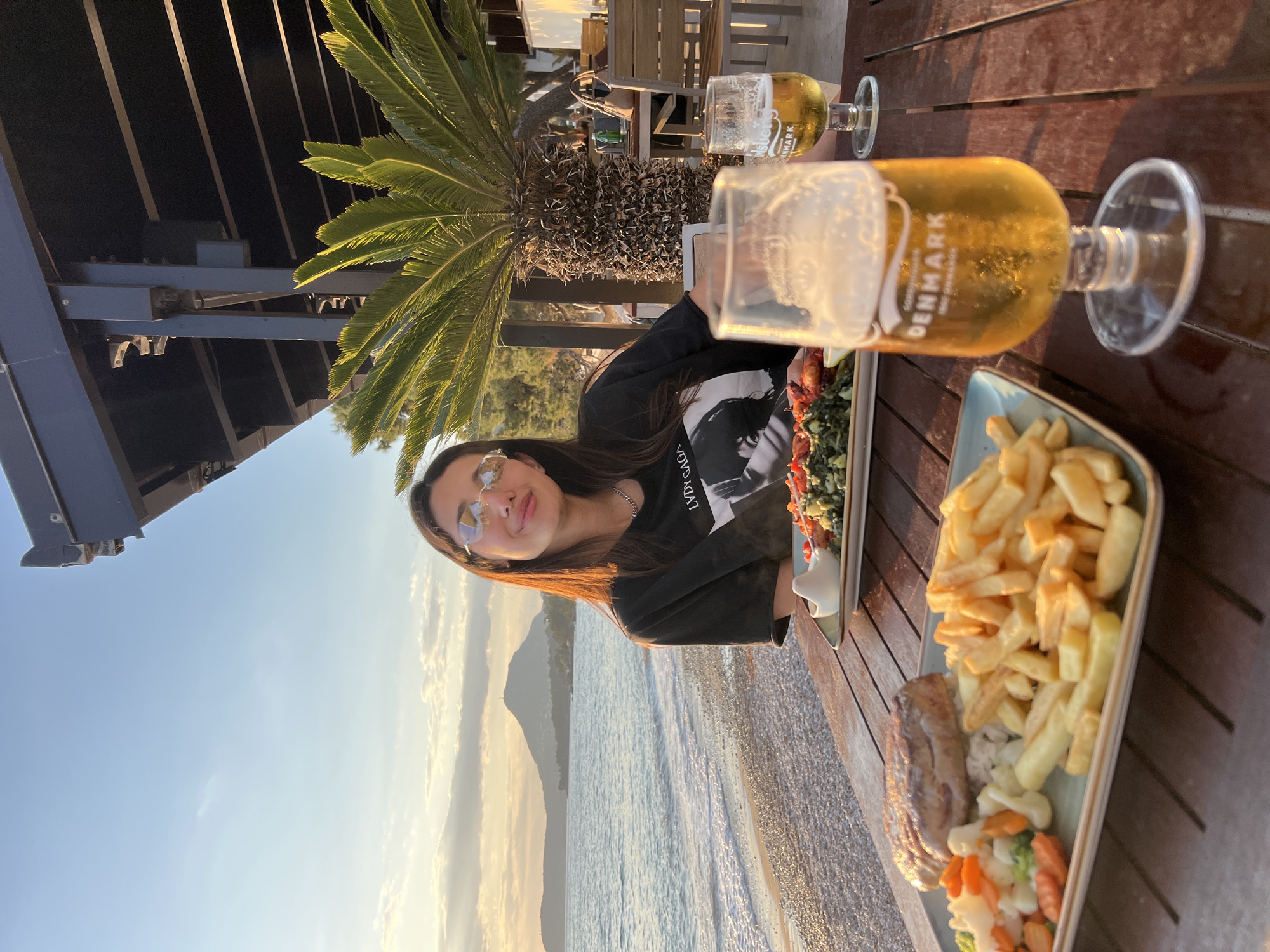 A woman is seated at a wooden table outdoors, with a partial view of a coastline at sunset in the background, likely in the city of Bar, Montenegro. She is wearing sunglasses and a dark shirt. On the table are two glasses of beer, and two plates of food, one with kebabs and greens and the other with fries, meat, and cooked vegetables. A palm tree is partially visible. The setting suggests a restaurant or bar near the sea.
