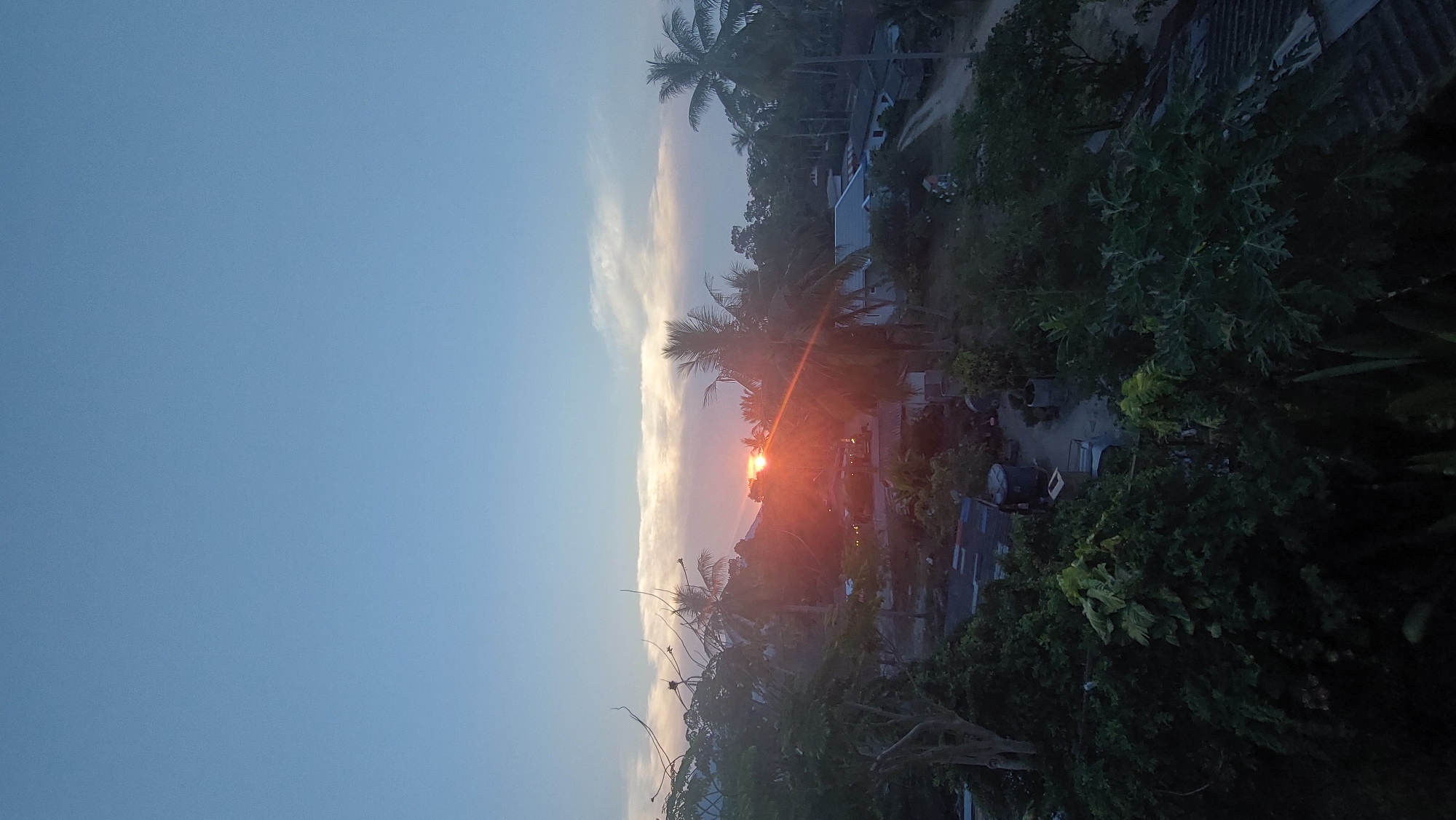 The image depicts a sunset in Baan Mae Nam, Thailand. The sky is a gradient of blue transitioning to orange and red near the horizon, where the sun is setting. Silhouetted palm trees are visible in the foreground against the bright light of the sunset. The sun's rays create flares across the scene. Houses and other buildings can be seen nestled amongst the trees, suggesting a residential area. The vegetation is lush and green. A dirt road can be discerned between the houses.
