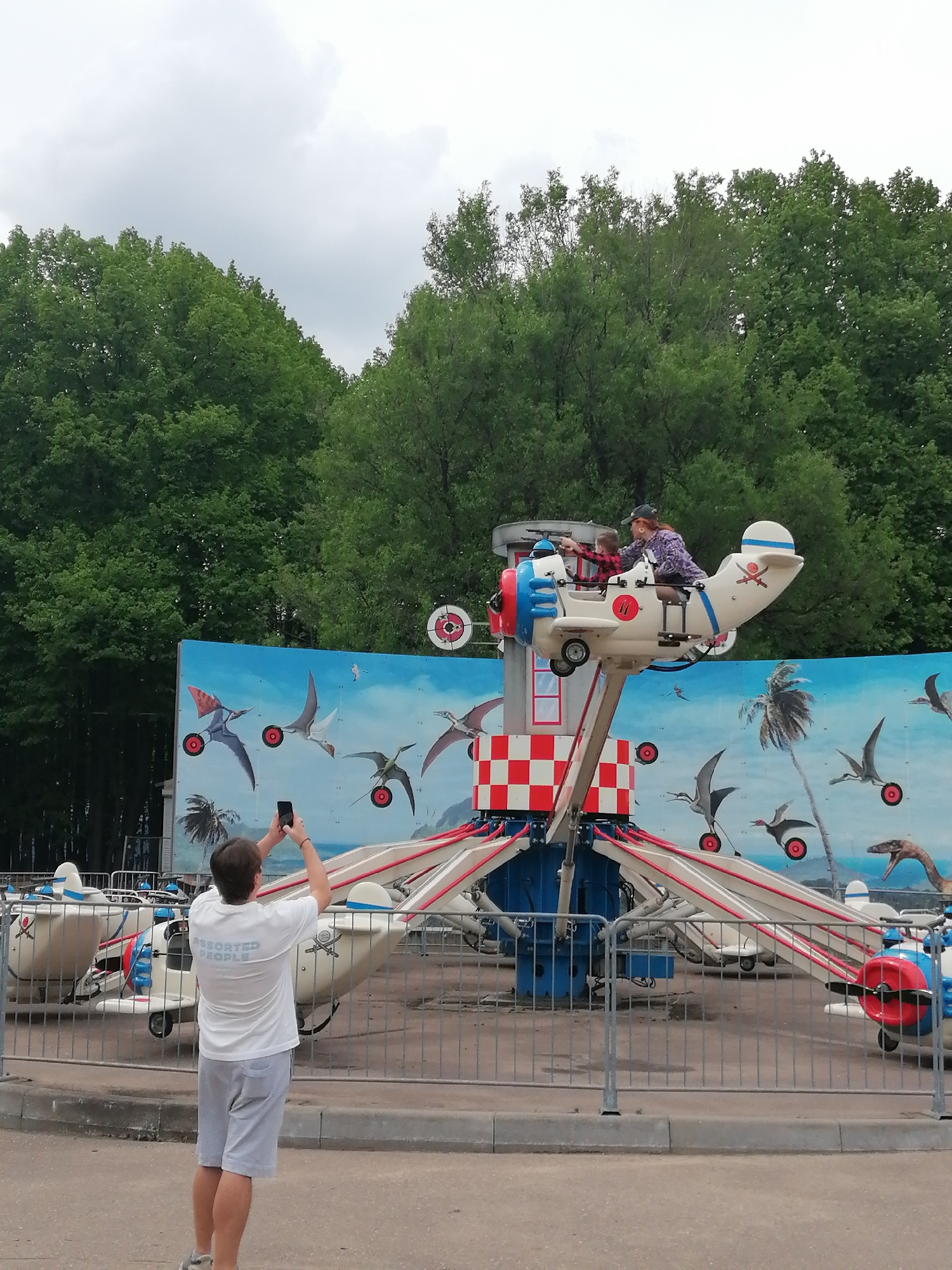 The image shows a scene at an amusement park in Moscow, Russia. A man stands in the foreground, facing away from the camera, holding a smartphone up to take a picture of a children's airplane ride. He is wearing a white T-shirt with "Assorted People" printed on the back, gray shorts, and sneakers. The airplane ride features small white planes with red and blue accents, designed to resemble vintage aircraft. Two individuals, an adult and a child, are riding in one of the planes. The background includes a blue mural with images of pterodactyls. The ride is surrounded by a metal fence, and behind the fence are green trees under a cloudy sky.
