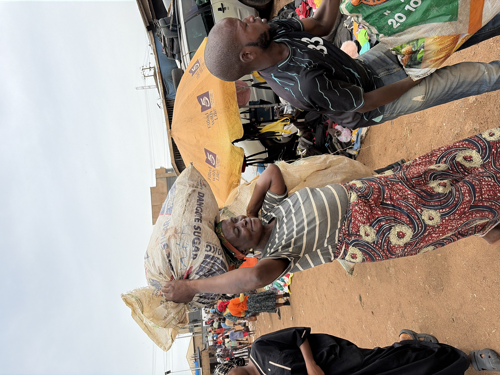In Gura Top, Nigeria, a woman wearing a striped gray shirt and a red and blue patterned skirt carries a large sack labeled "DANGOTE SUGAR" on her head. Another man stands nearby, carrying a bag marked with "20 10". An orange umbrella is nearby with the text "Filled with Life". The ground is dirt. A crowd of people is visible in the background.