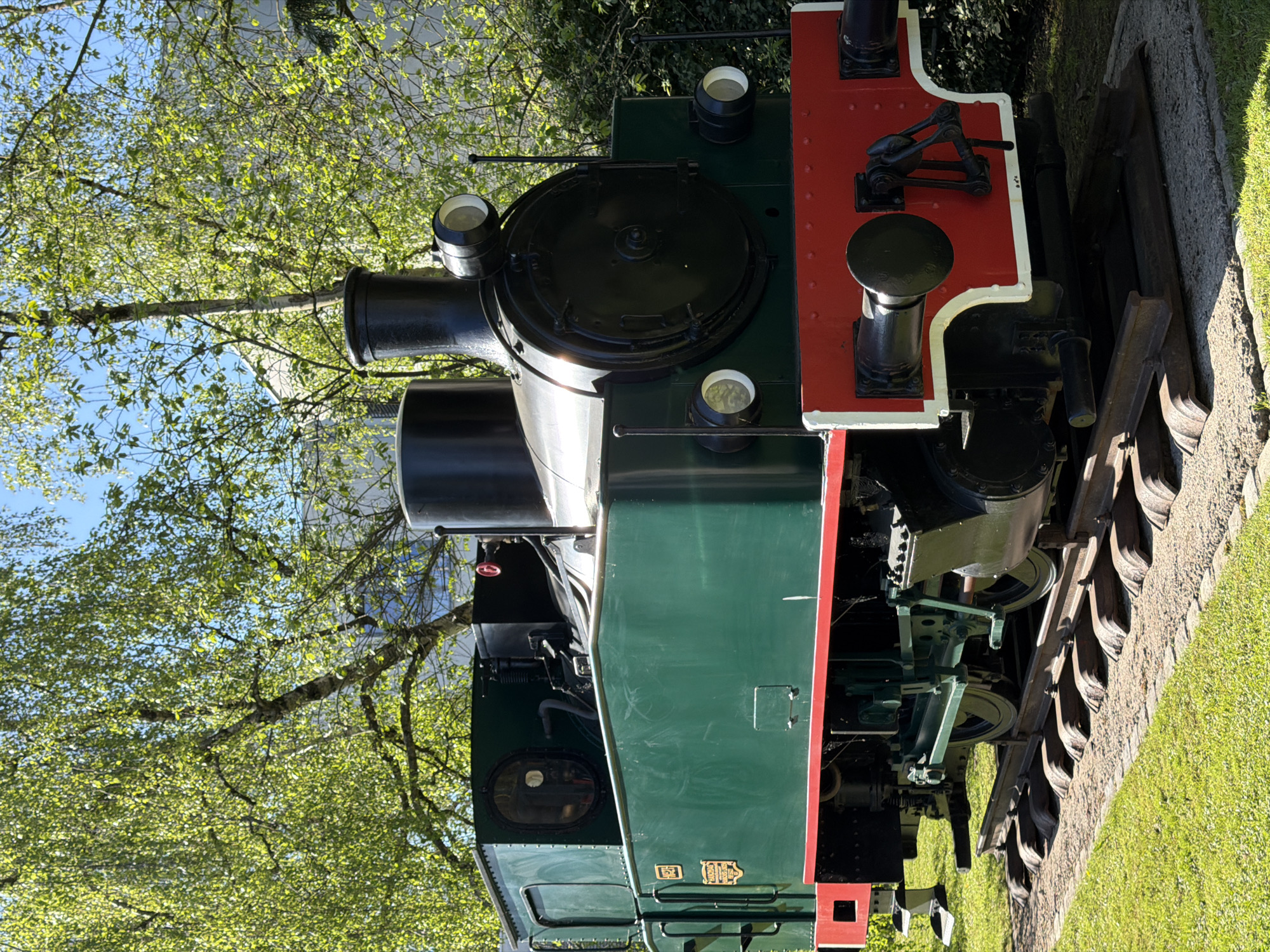 The image depicts a close-up view of an antique locomotive engine displayed outdoors, likely as a museum piece. The engine features a predominantly green body with red and white accents on its front section. Notable components include a large, circular black boiler top, several smaller cylindrical tanks or fittings, and complex mechanical linkages underneath. The engine rests on railway tracks embedded in a gravel path, with green grass visible alongside.
In the background, lush green trees with light-colored leaves are present, suggesting a park or garden setting. The location is Esch-sur-Alzette, Luxembourg. The image captures the details of the locomotive's construction and its weathered appearance in an outdoor environment.