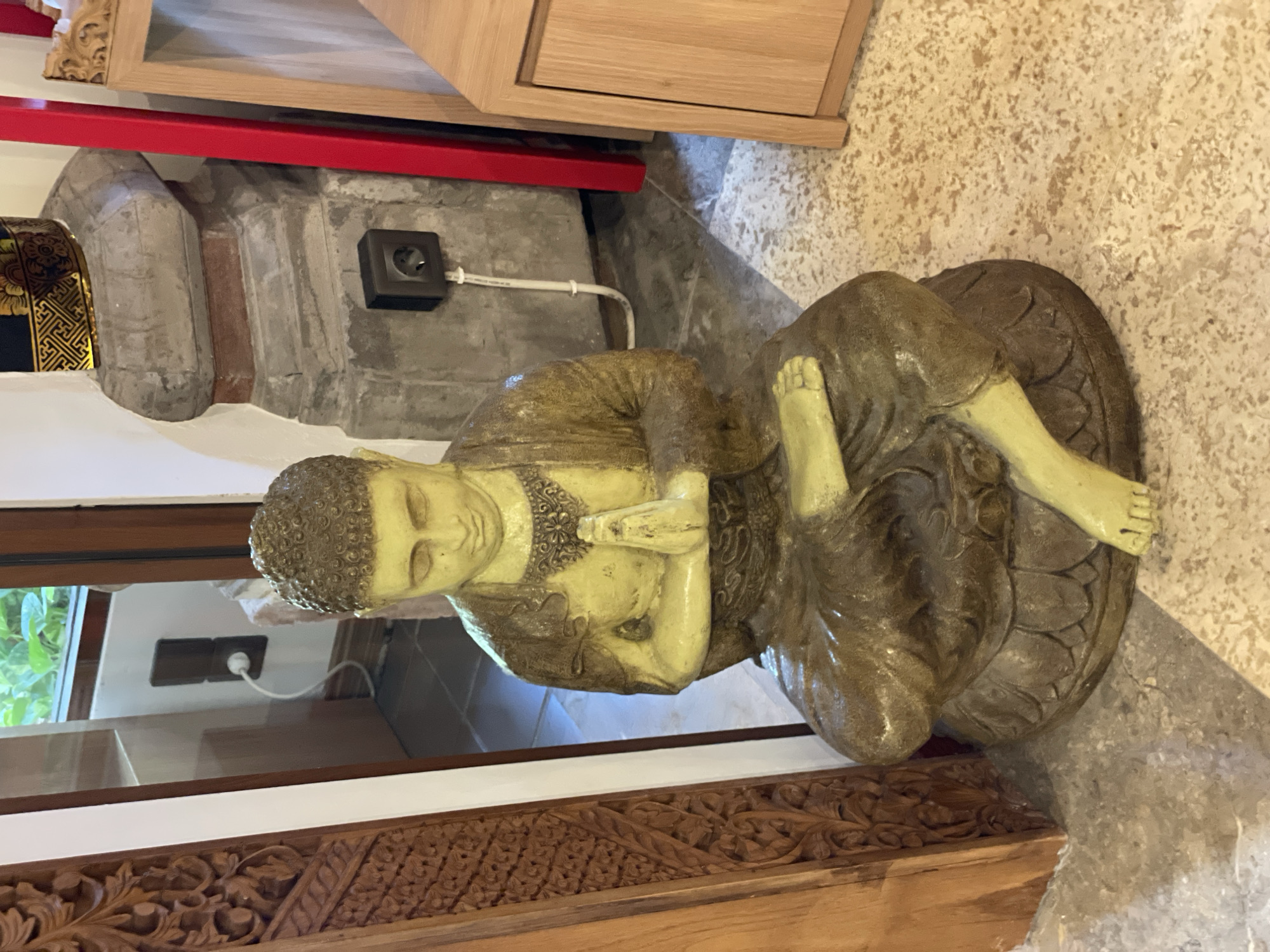 A statue of Buddha is seated in meditation on a lotus base. The statue is colored yellow and brown and is adorned with a necklace. The Buddha is positioned in front of a stone pillar with a red vertical beam and a wooden cabinet to the right. To the left of the Buddha statue is a carved wooden panel. The floor is a light-colored tile with a speckled pattern. There is an electrical outlet with a white cord plugged into it on the stone pillar. The background is blurred, but appears to be an indoor setting with plants visible through a doorway.