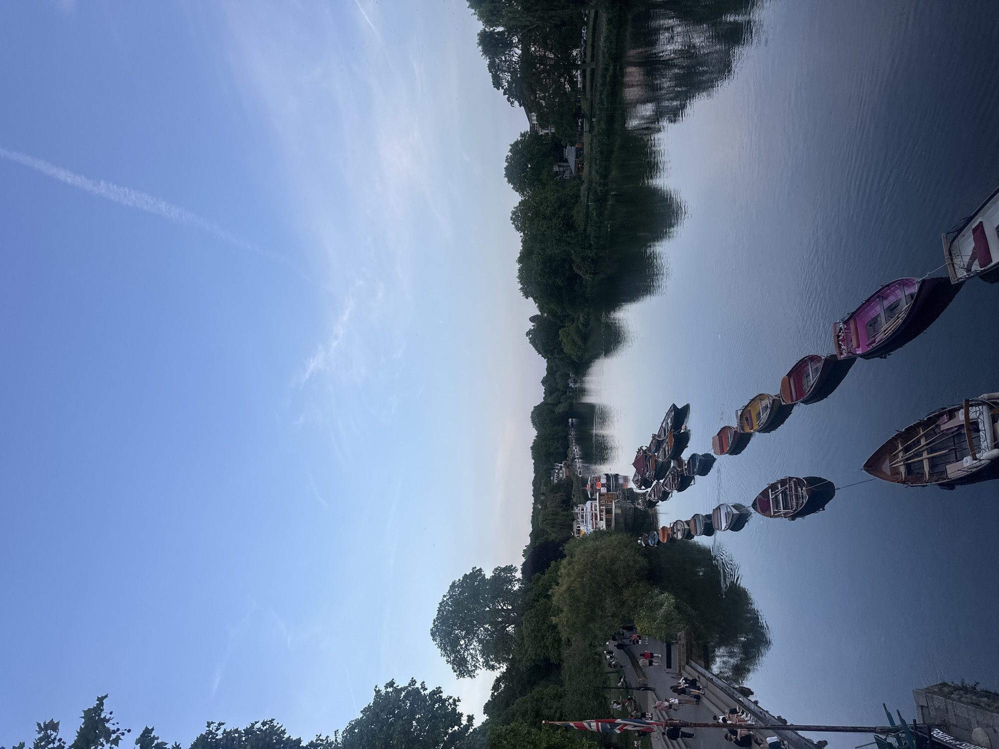 The image captures a serene riverside scene, likely along the River Thames in London, United Kingdom. A row of colorful boats, possibly for hire, is tethered to the riverbank. Trees and buildings line the opposite bank, reflected in the still water. A path runs alongside the river on the near side, populated with people walking and standing. The sky is a gradient of blue, suggesting twilight. A Union Jack flag is visible near the lower edge.
