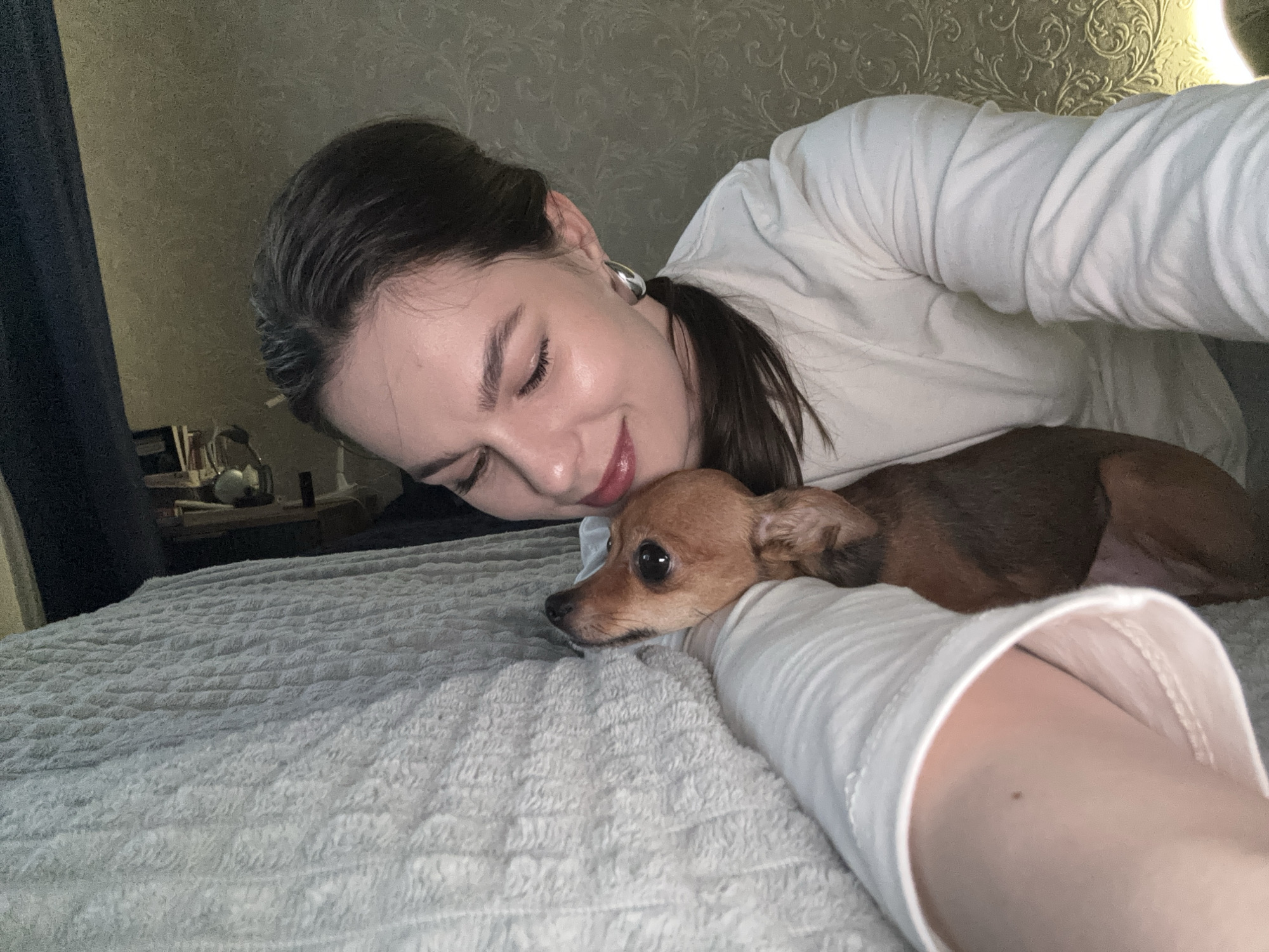 The image captures a tender moment between a woman and a small dog on a bed in what appears to be a cozy bedroom in Chaikovsky, Russia. The woman, with dark hair pulled back and wearing a white long-sleeved top, is seen from a close-up, slightly elevated perspective, suggesting a selfie or self-portrait. She lies on her left side, looking down with a gentle, contented smile, her eyes partly closed. A silver hoop earring adorns her left ear, and she has subtle makeup with dark lipstick.
Beside her, a small, reddish-brown dog, likely a Chihuahua or similar toy breed, rests calmly. The dog is positioned with its head near the woman's arm, gazing towards the left with soft, wide eyes. Both subjects appear relaxed and comfortable.
The bed is covered with a light-colored, textured blanket or comforter. The background reveals a wall adorned with elegant, patterned wallpaper, possibly a damask design. A soft, warm light emanates from the upper right, hinting at a lamp or other indirect light source, contributing to the subdued, intimate atmosphere. To the far left, a dark curtain or fabric is visible, and on a surface behind the woman, indistinct objects can be seen, possibly on a bedside table. The low lighting suggests it is evening or a dimly lit indoor setting. The overall mood conveys affection, comfort, and peaceful companionship. No visible text is present in the image.