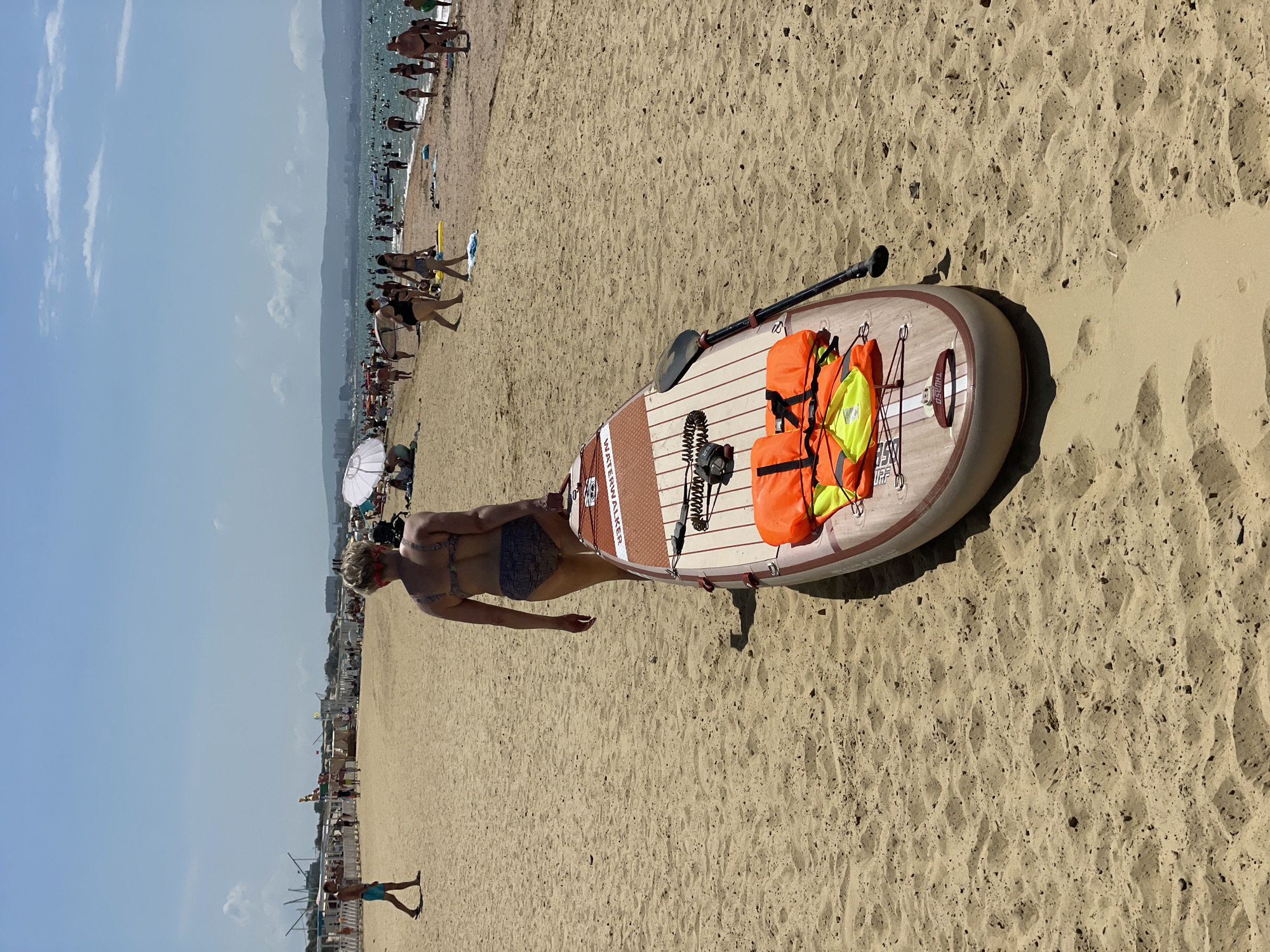 In Anapa, Russia, on a sunny day at the beach, a woman in a bikini walks along the sandy shore carrying a paddleboard. The paddleboard is light brown with red and white stripes and has two orange life vests and a black paddle attached. In the background, a crowded beach stretches out with people swimming in the sea, sunbathing on the sand, and walking along the water. Beach umbrellas and white structures are visible along the shoreline, and mountains can be seen in the distance. The sky is blue with scattered white clouds.