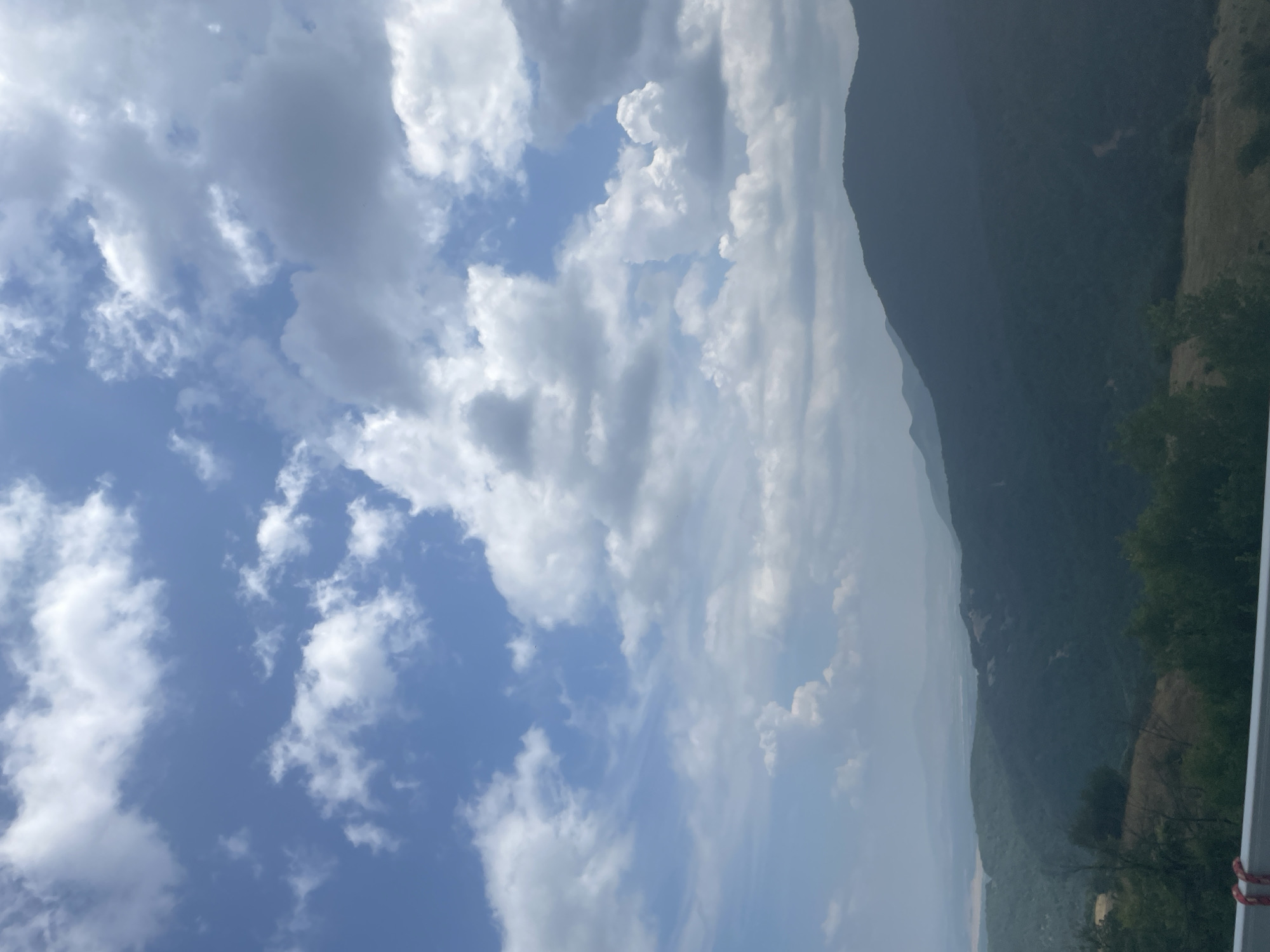 The image depicts a panoramic view of rolling hills and mountains under a partially cloudy sky. In the foreground, a white barrier with red and white rope is visible, suggesting a viewpoint or a roadside. The landscape is characterized by dense, dark green foliage covering the slopes of the hills, with some patches of lighter vegetation and bare earth visible. In the distance, layers of haze obscure further mountain ranges, creating a sense of depth. The sky is a bright blue with scattered cumulus clouds, some of which are large and shadowed, indicating potential for changing weather. The overall scene is natural and undeveloped, evoking a sense of natural beauty and vastness. The location is identified as Oreshets, Bulgaria. There are no people or specific actions depicted in the image.