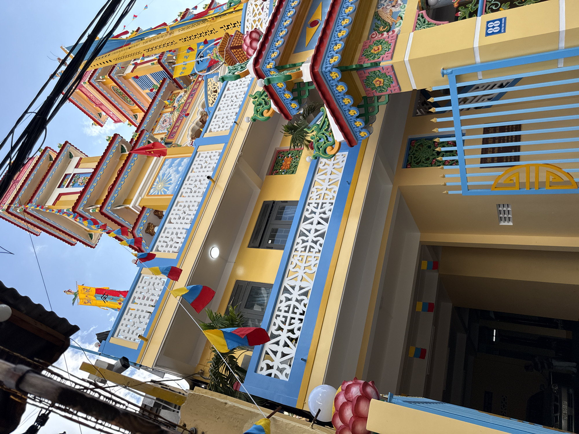 The image shows a brightly colored, multi-story building with ornate architectural features, likely a temple or pagoda, situated in Nha Trang, Vietnam. The building is painted predominantly in yellow and blue, with red accents on the roofs and decorative elements. Flags in red, yellow, and blue are hung across the facade. The structure has multiple levels with balconies and windows. Decorative motifs, including what appear to be dragons and floral patterns, are visible. In the foreground, a blue metal gate with a yellow circular design is present. A license plate is visible on a parked car under an overhang on the left side of the image. The sky above is blue with some scattered clouds. The overall impression is of a vibrant and culturally rich religious or community building in an urban Vietnamese setting.