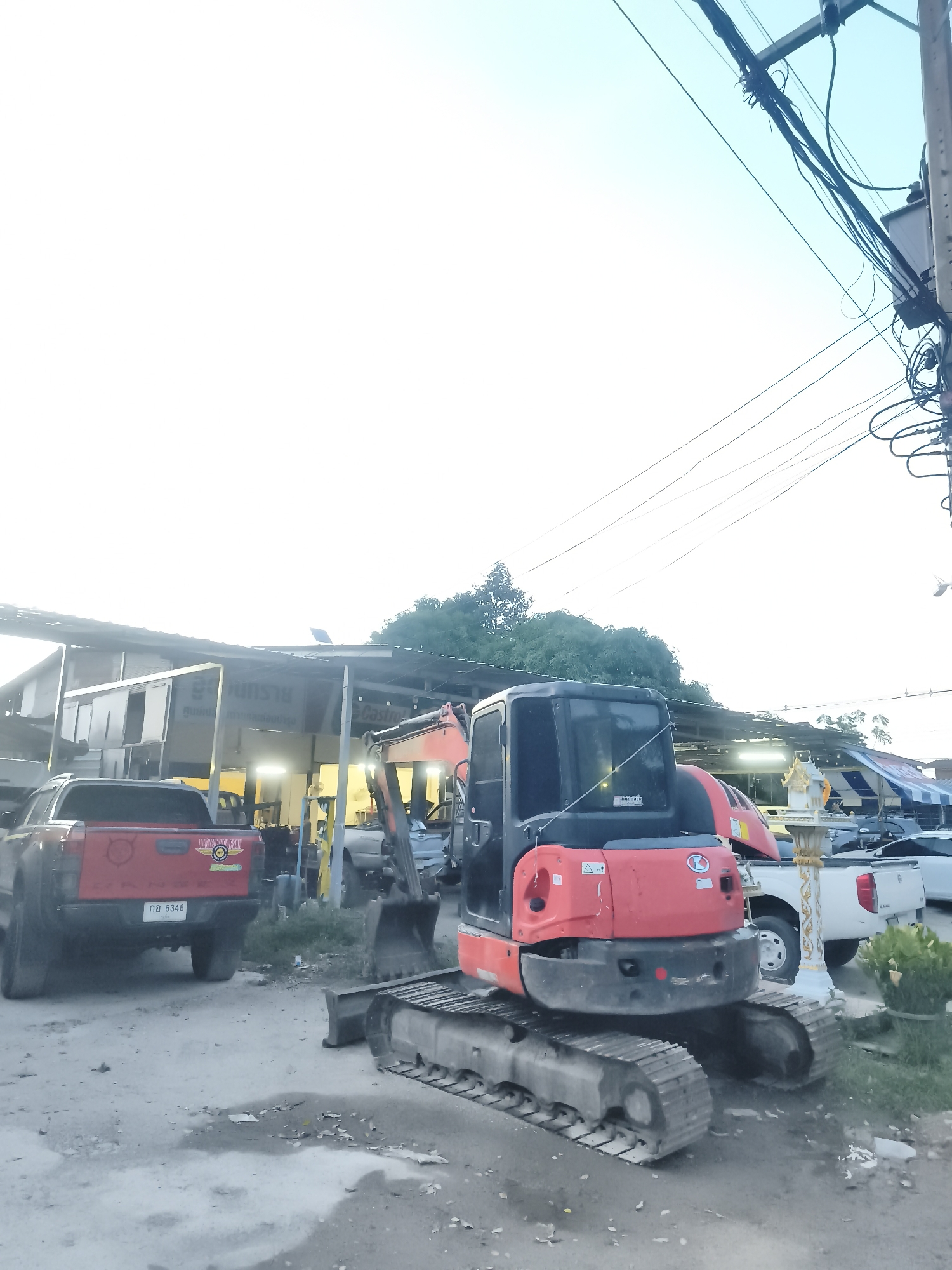 The image shows a compact excavator parked on a paved surface. Behind the excavator, a red pickup truck is visible. Further back, there is a building with open bays. Other vehicles are parked near the building. Overhead, power lines run along poles. The location is identified as Baan Mae Nam, Thailand.