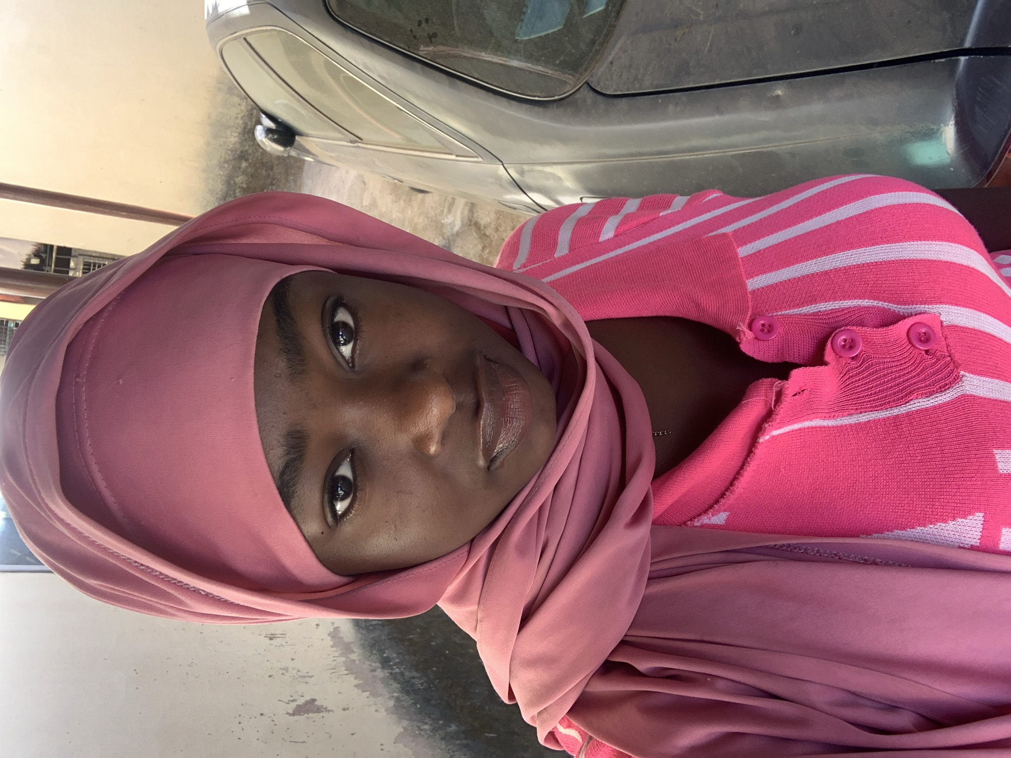 A medium shot depicts a dark-skinned woman wearing a pink headscarf and a pink and white striped cardigan. She is positioned in front of a silver car, potentially within an urban environment. The location is identified as Rafin Par, Nigeria. No specific actions or interactions are visible.