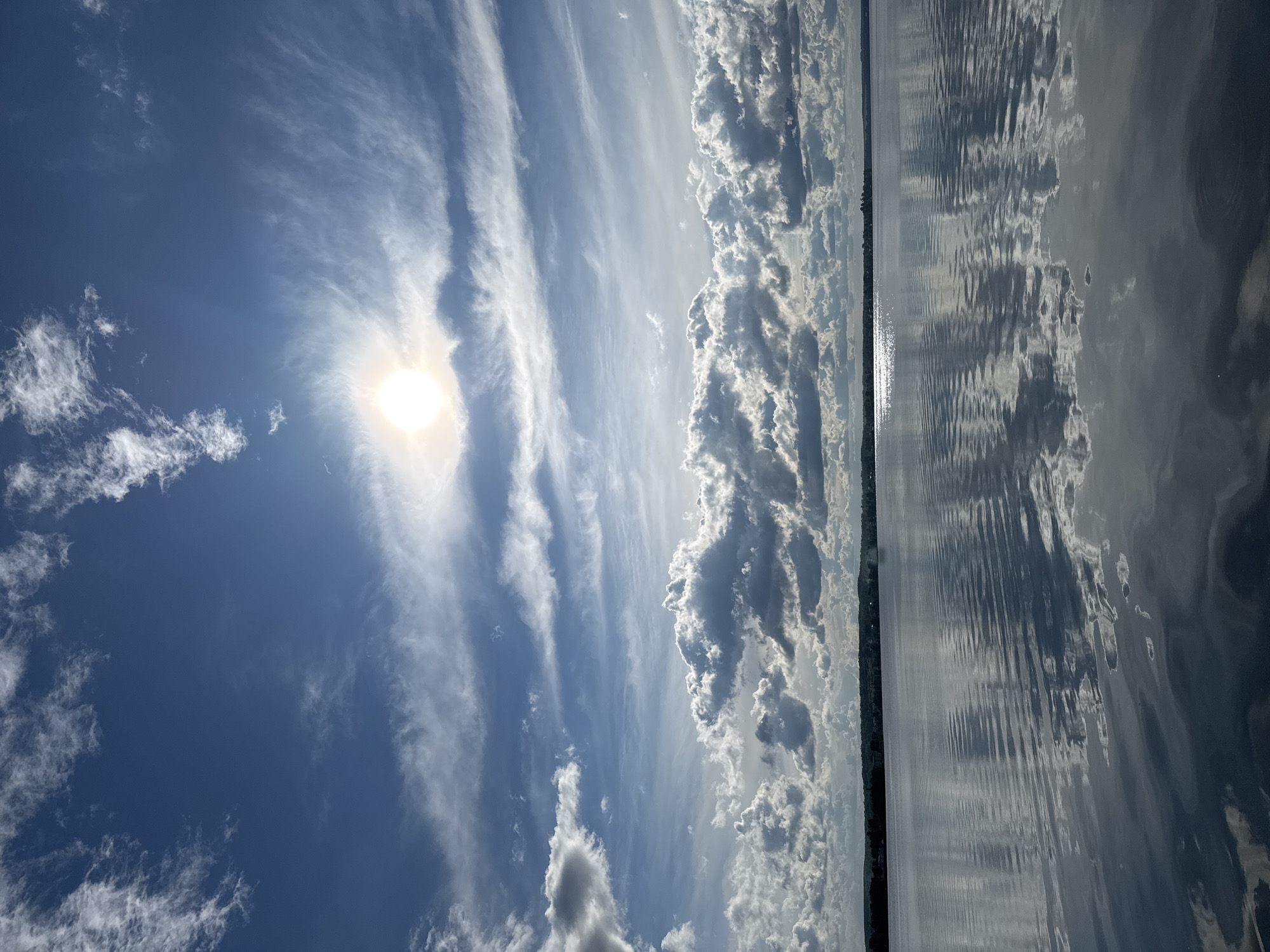 The image shows a body of water meeting the sky. The left portion of the image depicts a bright blue sky with scattered white clouds and the sun. The right portion depicts water reflecting the sky. A dark strip of land, potentially a shoreline, divides the sky from the water. The location is likely Votkinsk, Russia. No people or specific objects are discernible.