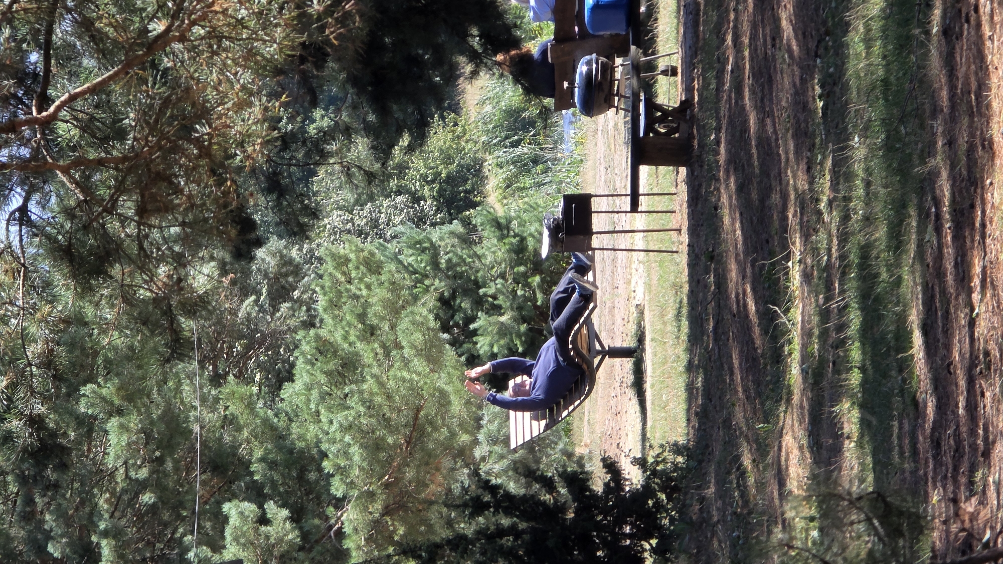 A person reclines on a wooden lounger with their arms raised, seemingly relaxed, in an outdoor setting with trees and grass. In the foreground, a barbecue grill stands on a metal stand. Further back, a picnic table is visible, and a few other people are seated on benches in the background. The scene appears to be a park or a recreational area.
