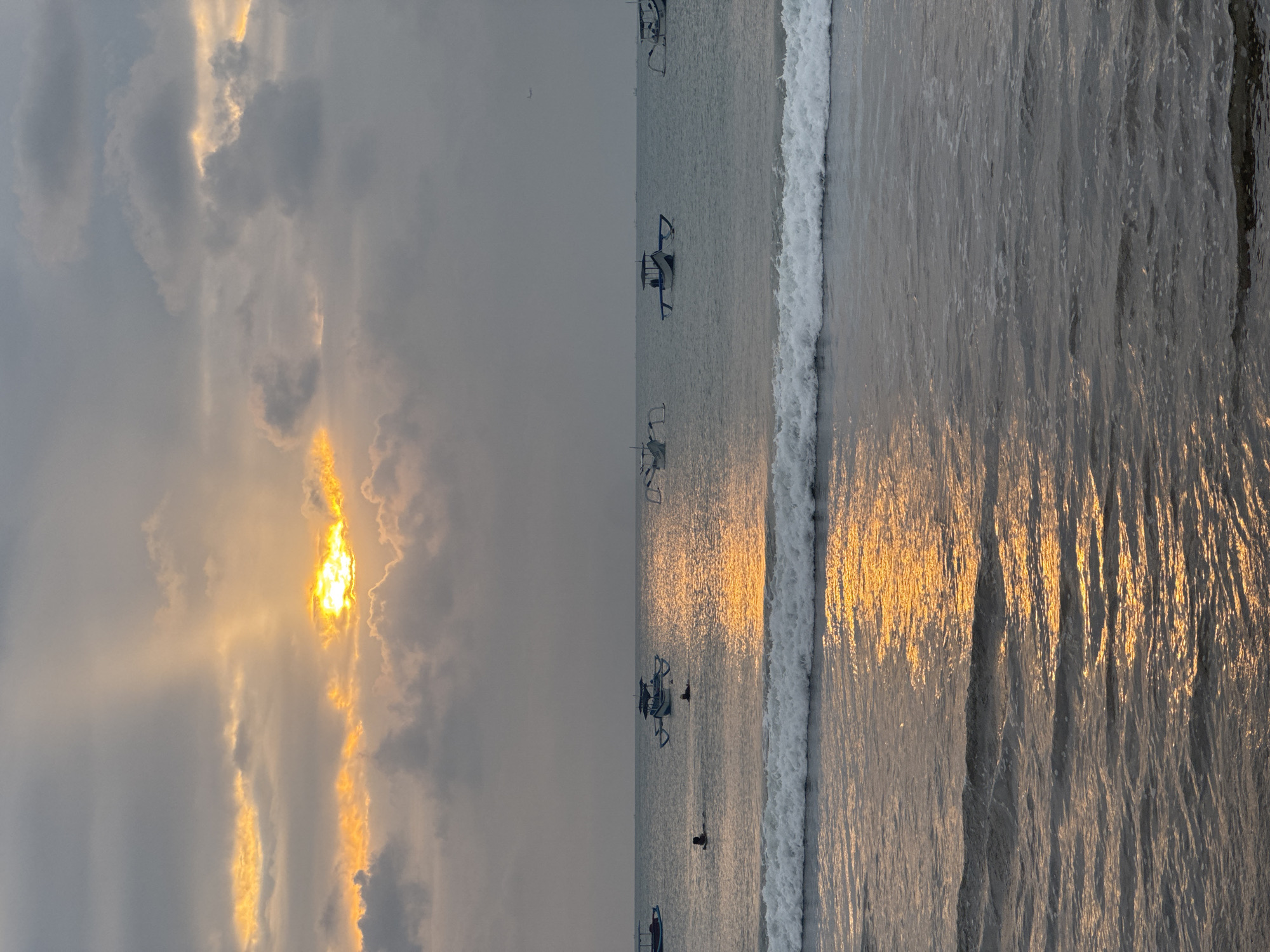 This is a vertical, wide-angle photograph of a beach scene at sunset. The left side of the image features a dramatic sky with the sun partially obscured by dark, textured clouds, allowing bright, golden rays to break through. The right side of the image shows the ocean with a breaking wave on the far right. The water reflects the golden light from the sunset, creating shimmering patterns on its surface. Several traditional outrigger boats are visible in the water, some with people on or near them. Two individuals are seen in the water, appearing to be swimming or wading. The overall mood is serene and tranquil, with the warm sunset colors contrasting with the cooler tones of the sky and ocean. The location appears to be a tropical beach, likely in Bali, Indonesia, given the presence of the outrigger boats commonly found there. The time of day is clearly sunset, indicated by the low angle of the sun and the golden light. The weather seems to be partly cloudy, with the clouds adding depth and drama to the sky. No visible text is present in the image.