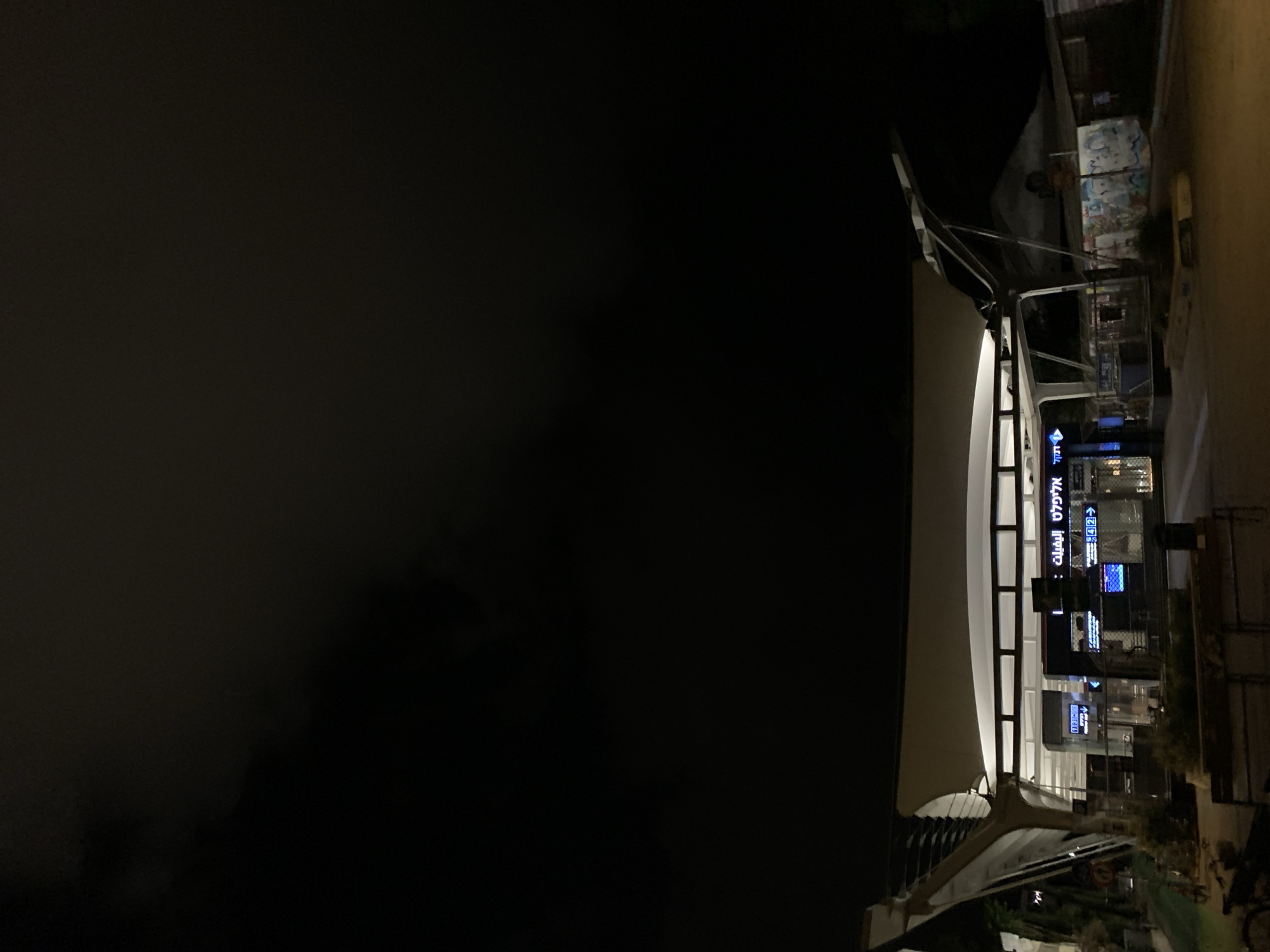 The image depicts a nighttime scene at Ben Gurion Airport in Tel Aviv, Israel. A covered walkway or bridge is visible on the right side of the image, featuring a series of glowing blue directional signs in both Hebrew and English. The signs likely indicate directions to various gates or services within the airport. The left side of the image is mostly dark, suggesting a nighttime sky or background. The walkway is lit from within, creating a contrast with the dark background. Outside the bridge there is a building with graffiti on it.
