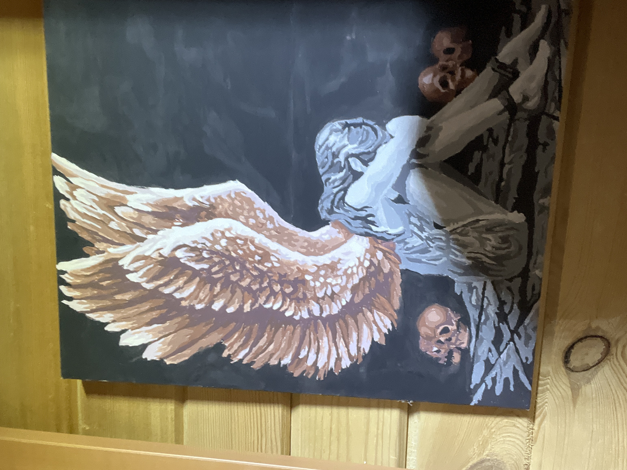 The image displays a painting featuring a figure hunched over with its head in its knees, partially obscured by large, feathered wings. The figure's skin appears pale, and its hair is depicted in shades of grey. Around the figure, several skulls are scattered on a dark, textured surface. The background is a deep, dark color, possibly black or dark gray. The painting is framed and hangs on a wooden wall. There is no information in the image to indicate a location in Ashkadar, Russia.