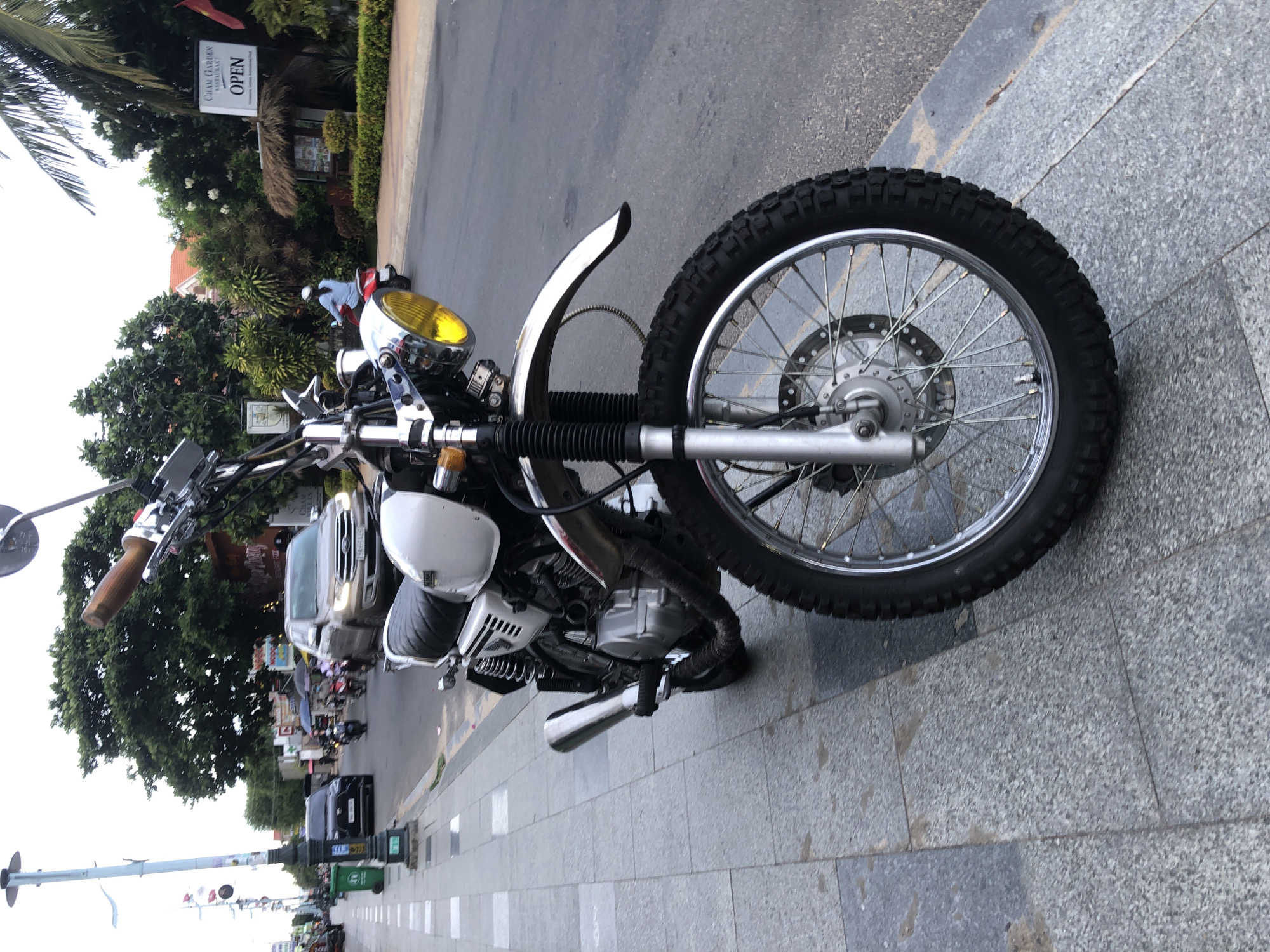 A white and silver motorcycle is parked on a gray, tiled sidewalk next to a street in Phu Thuy Ward, Vietnam. The motorcycle is facing the viewer, with its front wheel and handlebars prominently displayed. The street is wet, suggesting it may have recently rained. In the background, trees and buildings line the street, and several cars and motorcycles are visible. A sign for "CHAM GARDEN RESTAURANT" with the word "OPEN" is visible on the right side of the image. The sky is overcast. The sidewalk has a green trash can on the left side.
