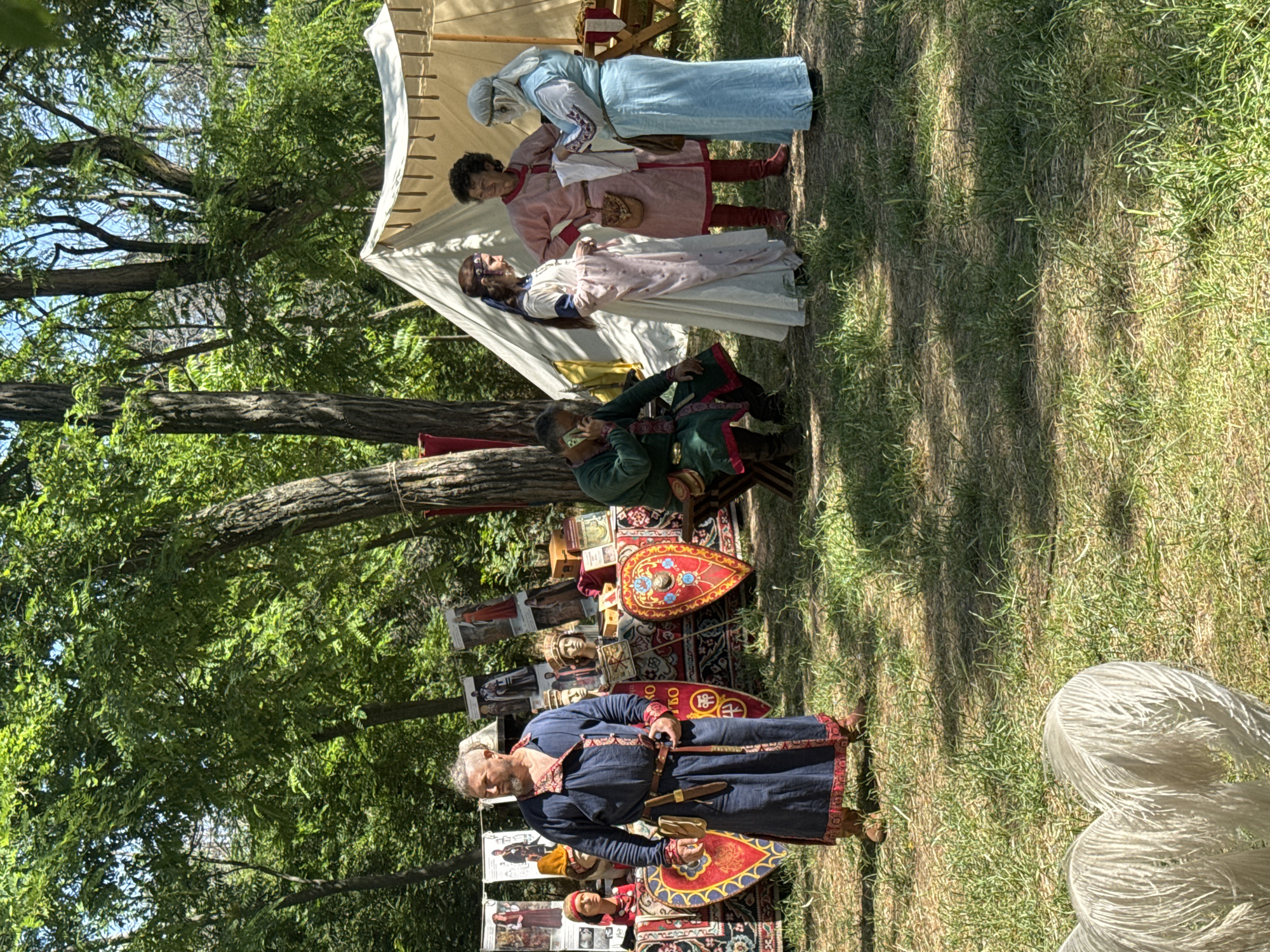 The image depicts a historical reenactment event taking place outdoors, likely in a park or open field, on a sunny day. In the foreground, a man in a long, dark blue robe with red trim stands looking to his left. He wears a belt with a dagger and a pouch. Behind him, a table displays various historical artifacts, including shields with intricate designs, mannequins with historical headwear, and framed images. To the right, a group of four people are engaged in conversation. Three individuals are dressed in historically inspired attire. A man in a green tunic is seated and appears to be on a phone. Three women, two in long dresses and one in a light blue robe, are standing and interacting. In the background, tents and trees are visible, suggesting a festival or fair. The overall scene conveys a sense of historical immersion and community gathering. This event appears to be held in Plovdiv, Bulgaria, possibly during a historical festival like "The European Capital of Culture Plovdiv 2019" or similar historical reenactment events.