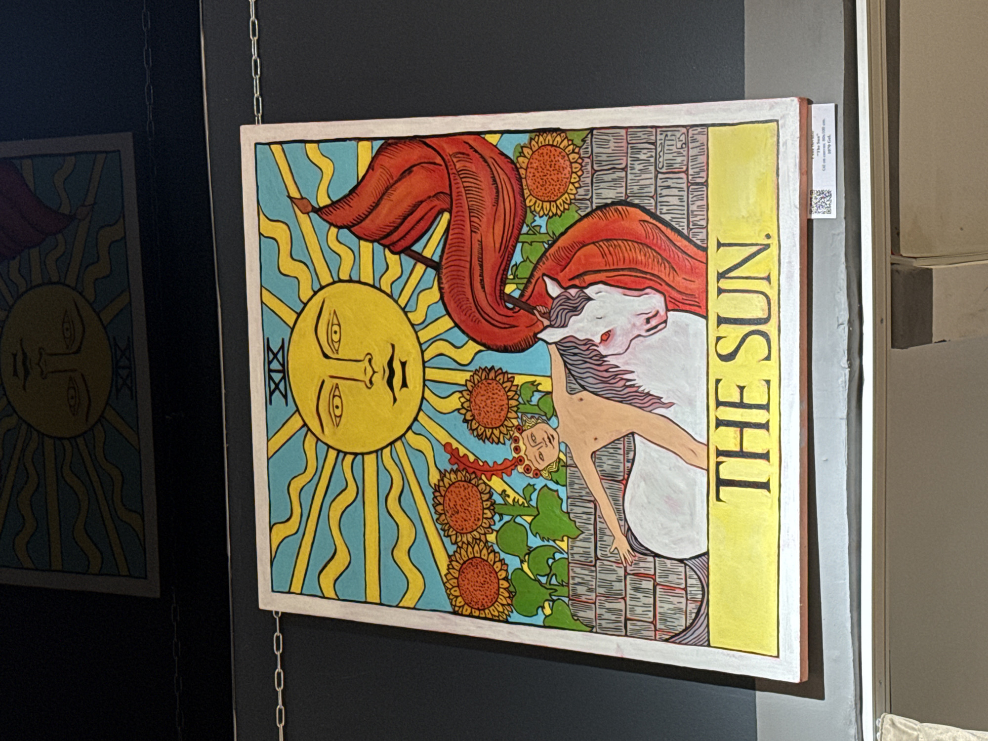 The image shows a painting of the "The Sun" Tarot card hanging on a wall. The painting is brightly colored and features a yellow sun with a human face, sunflowers, a naked child riding a white horse, and a red banner. The words "THE SUN" are printed vertically along the right side of the painting. Another blurry painting of the sun can be seen in the reflection. There is a tag attached to the side of the painting with additional information, possibly including a QR code. The setting appears to be an art gallery or exhibit, likely in Batumi, Georgia, based on the request context.
