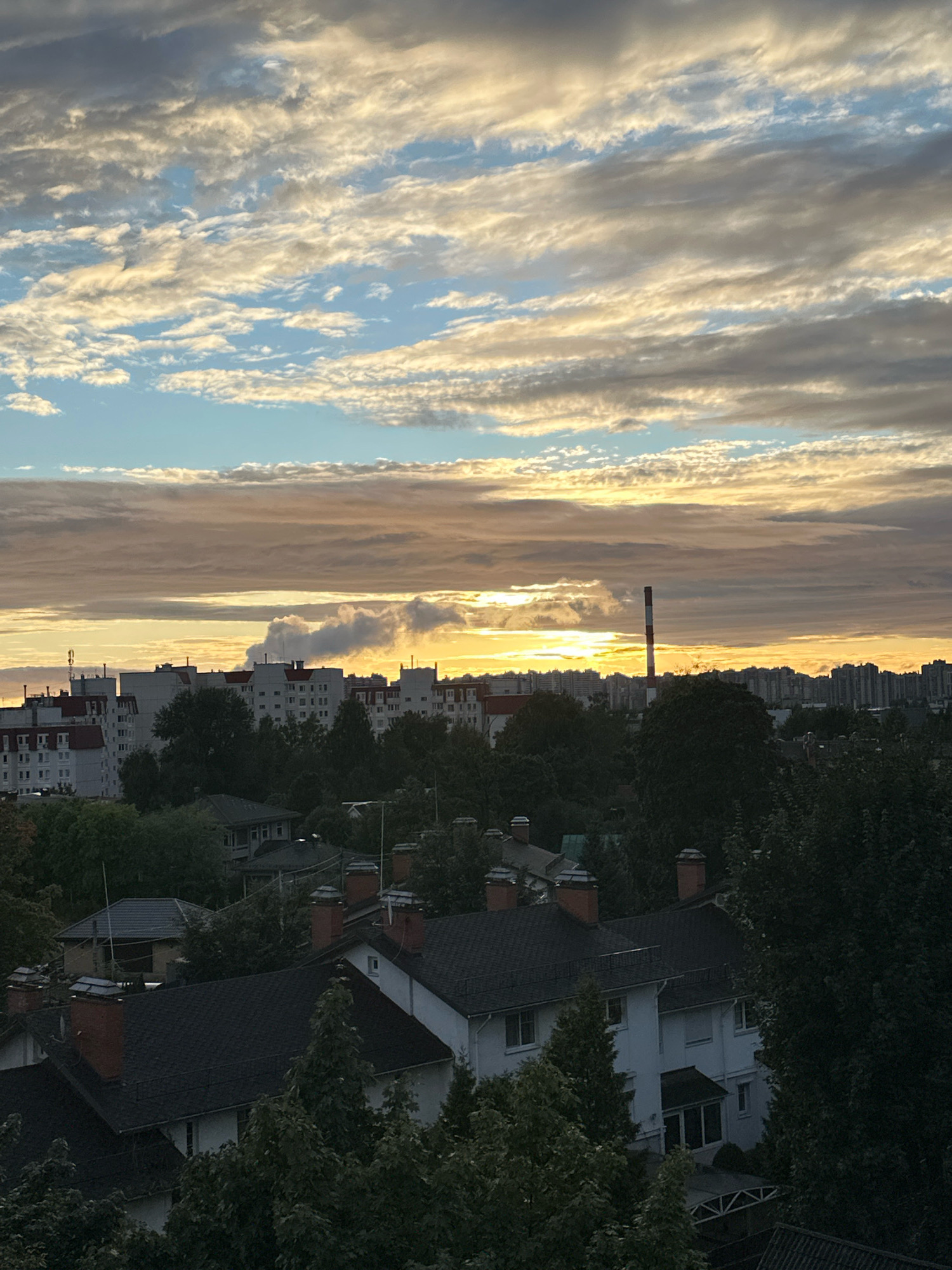 The image captures a view of a city skyline at dawn or dusk. The sky is filled with a mix of clouds, with hues of orange, yellow, and blue suggesting the low angle of the sun. In the middle ground, there are several apartment buildings, some with white facades and red-tiled roofs. A tall industrial chimney with red and white bands stands prominently against the sky, emitting a plume of white smoke. In the foreground, the tops of trees and residential houses with dark roofs and chimneys are visible. The overall scene depicts a residential and industrial area under a dramatic sky.