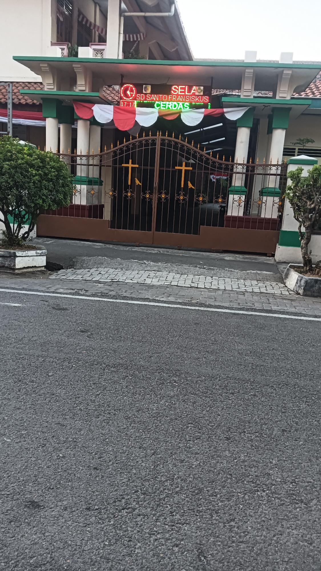 This image captures the entrance to SD Santo Fransiskus in Sragen, Indonesia. The school's name is displayed on a digital LED sign above a pair of ornate brown metal gates. The sign also shows the time "17:17" and the word "CERDAS" in green. Red and white banners are draped across the top of the entrance, and a green and white striped canopy extends over the gate. The gates themselves are decorated with gold crosses and bird figures. To the left of the gate is a potted green shrub, and to the right is a small tree. The school building's facade, with its tiled roof and white and green accents, is visible behind the entrance. The ground in front of the gate is paved with cobblestones, leading to an asphalt road. The overall scene depicts the main entrance of a school, likely at the end of the school day given the time displayed.