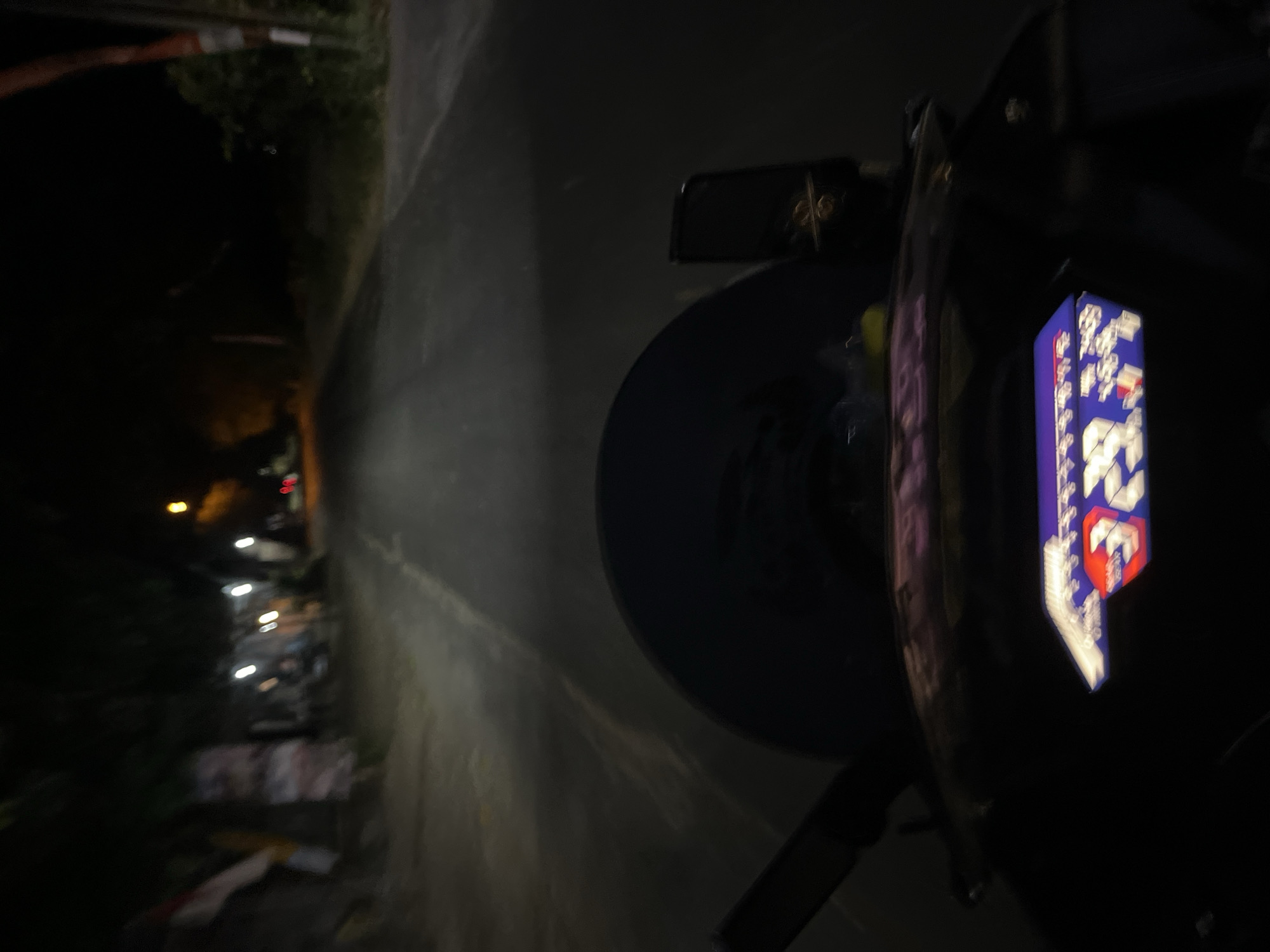 The image is taken from the perspective of someone riding a motorcycle at night in Pecangaan, Indonesia. The road is dimly lit, with streetlights visible in the distance. The motorcycle's dashboard is illuminated, displaying a digital readout with numbers and symbols. The surroundings are dark, with trees and buildings lining the road. The scene suggests a quiet, rural setting within the city.