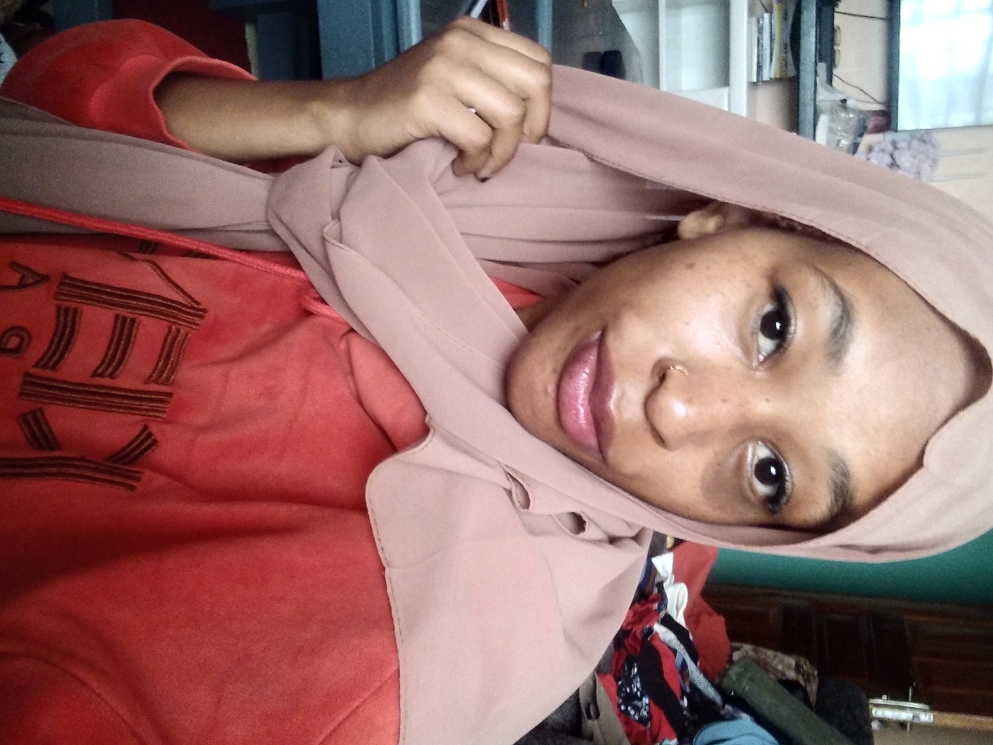 A young woman is pictured in a close-up, head-and-shoulders shot, wearing a light pink hijab and a salmon-colored hooded sweatshirt. She has dark skin, dark eyes, and a small nose ring. Her lips are painted with a glossy pink lipstick. She is gently touching her hijab with her right hand. The background is blurred, but appears to be an indoor setting with shelves and clothing visible. The image is taken in Nyeri, Kenya.