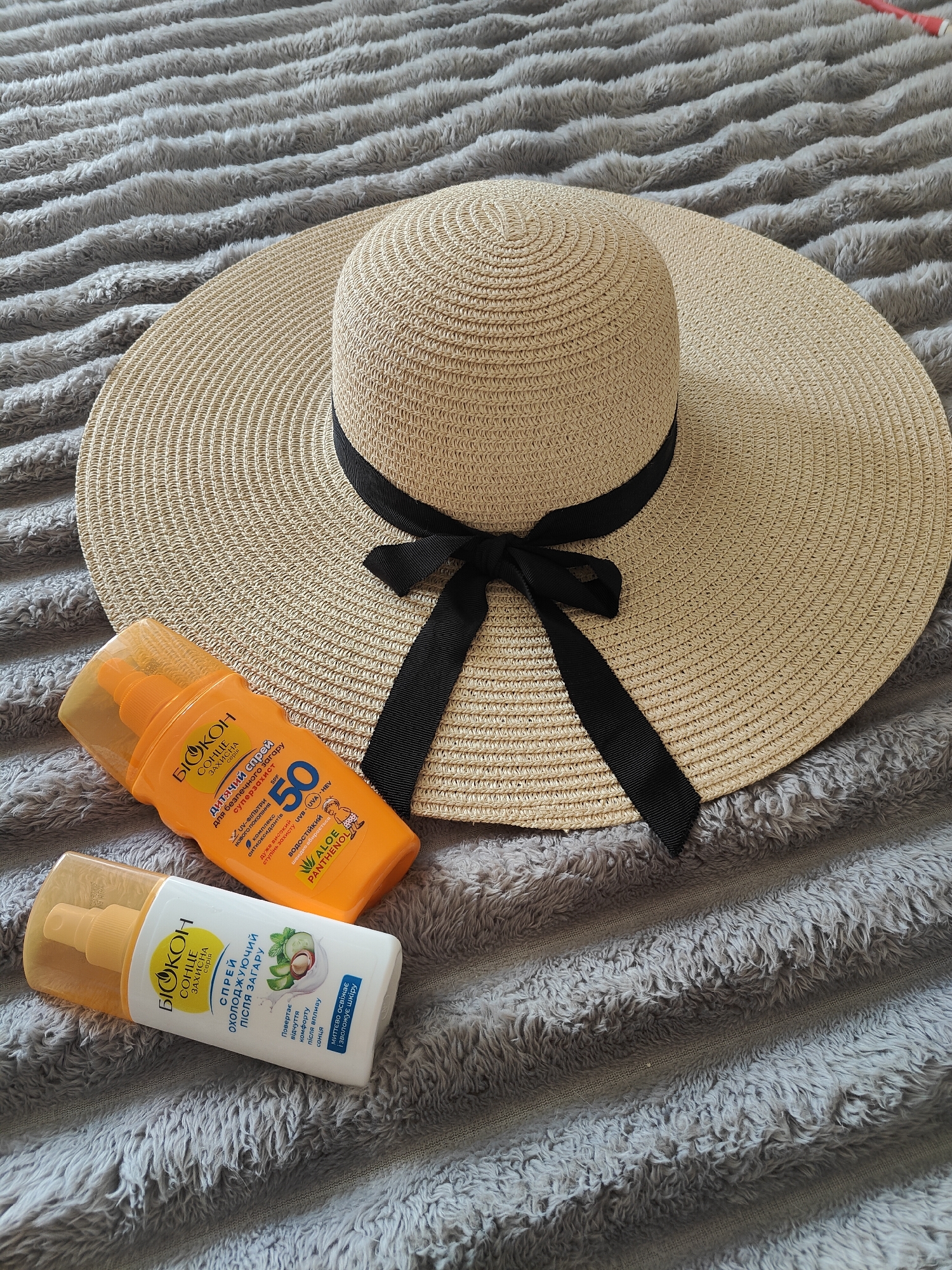 This media file displays a flat lay composition featuring sun protection essentials. The central object is a wide-brimmed straw hat with a black fabric ribbon tied into a bow, positioned slightly off-center towards the top and right. To the lower left of the hat are two bottles of cosmetic products, arranged diagonally.
The items are placed on a soft, grey, textured blanket or throw, which features prominent horizontal ridges, providing a tactile background.
The orange bottle, a sunscreen spray, is labeled "БІОКОН СОНЦЕ ЗАХИСНА серія" (BIOCON SUN PROTECTIVE series) and "дитячий спрей для безпечного загару" (children's spray for safe tan). It boasts "суперзахист" (super protection) with SPF 50, indicating "дуже високий ступінь захисту" (very high degree of protection) against "UVB (UVA)-HEV" rays, utilizing "UV-ФІЛЬТРИ НОВОГО ПОКОЛІННЯ" (new generation UV filters) and an "комплекс антиоксидантів" (antioxidant complex). It is also described as "ВОДОСТІЙКИЙ" (waterproof) and "швидко вбирається" (quickly absorbed), containing "ALOE PANTHENOL".
The white bottle, an after-sun spray, is also from "БІОКОН СОНЦЕ ЗАХИСНА серія" and is identified as "СПРЕЙ ОХОЛОДЖУЮЧИЙ ПІСЛЯ ЗАГАРУ" (cooling after-sun spray). Its text promises to "Повертає відчуття комфорту після впливу сонця" (Restores feeling of comfort after sun exposure) and "миттєво освіжає і зволожує шкіру" (instantly refreshes and moisturizes skin). An illustration of cucumber slices and mint is visible on its label.
The scene suggests preparation for a sunny day, likely a vacation or beach outing in a warm climate, fitting for a coastal city like Chornomorsk, Ukraine. The items convey a sense of care for skin health and comfort during sun exposure. There are no people visible, and the time of day is not explicitly shown but implied to be when the sun is out.
