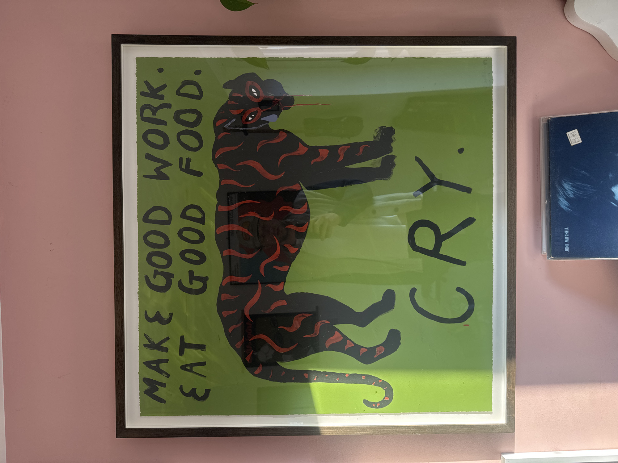 An interior shot features a framed artwork mounted on a light pink wall. The artwork displays a highly stylized, black, tiger-like creature with striking red wavy markings, set against a vivid lime-green background. The creature is depicted vertically, with multiple white eyes and a long, pointed snout from which a red line protrudes. Its front paws are extended forward, and it has a long, curled tail.
Bold black capitalized text is integrated into the artwork. On the left side, reading vertically upwards from bottom to top, it says: "MAKE GOOD EAT GOOD" followed by "WORK. FOOD." On the right side, also reading vertically upwards, is the word "CRY." The artwork is presented within a dark wooden frame, bordered by a crisp white mat.
To the right of the framed piece, partially visible against the pink wall, is a blue record album cover. A white sticker with "£5.00" and other faint details is attached to the cover, below which the artist's name, "JONI MITCHELL," is legible. A fragment of a green plant leaf can be seen at the top edge of the image, just above the artwork. Reflections on the artwork's glass suggest an indoor setting illuminated by natural daylight. The scene captures a static display of personal decor.