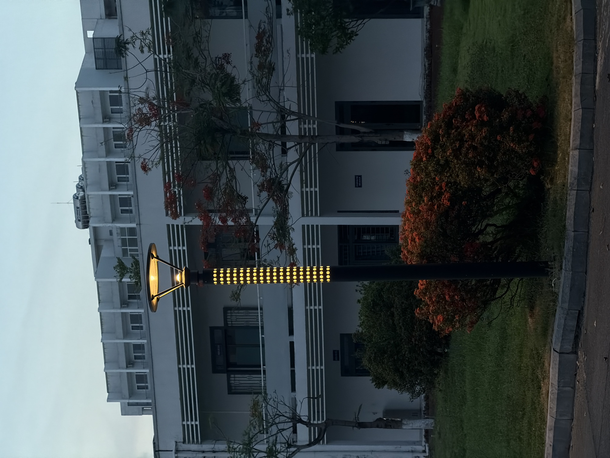 The image shows a multi-story building in what appears to be Đà Nẵng, Vietnam. A modern streetlight stands in the foreground. The streetlight is black with yellow circular lights arranged in rows along the pole. A red-flowered tree is situated next to the streetlight. The building is white with dark window frames. There are trees and grass in the area around the building. A sidewalk borders the grassy area. The sky is overcast. The image is rotated counter-clockwise.