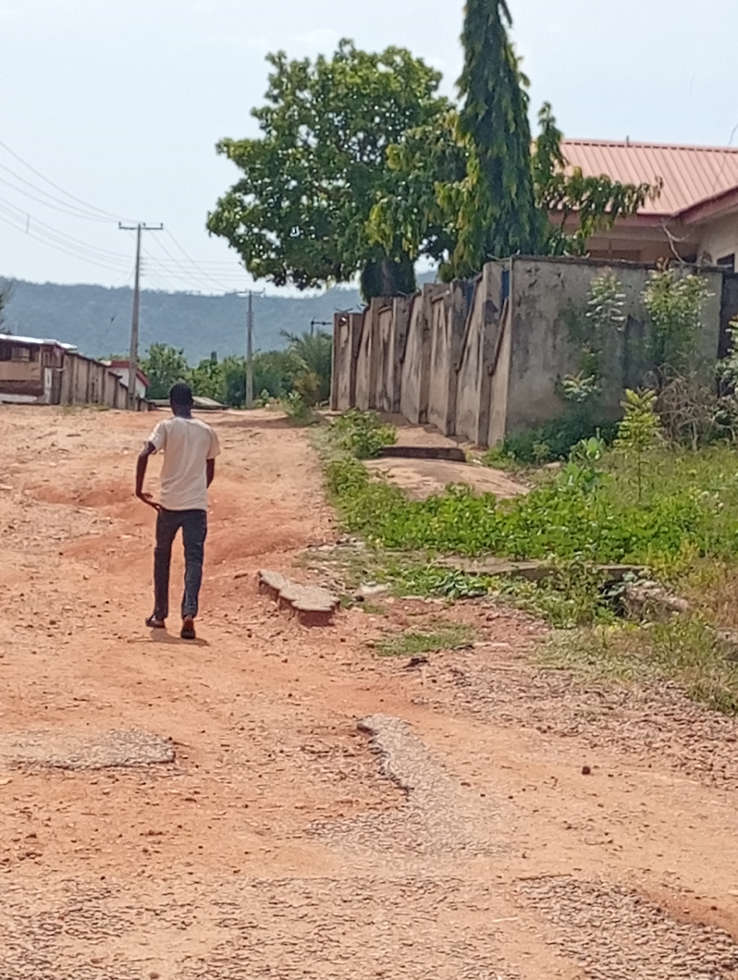 A person is walking on an unpaved road in Lokoja, Nigeria. The individual is wearing a light-colored t-shirt, dark pants, and dark sandals. They are walking away from the viewer, down a road that has a mixture of dirt, gravel, and patches of concrete. The road appears to be in disrepair with cracks and eroded areas. On the right side of the road is a concrete wall, with greenery and vegetation growing in front of it. On the left side of the road are buildings and structures. In the background, there is a distant view of a hill or mountain. Overhead power lines and utility poles are visible. A tree is partially blocking view in the background and to the right is a house with a red roof.