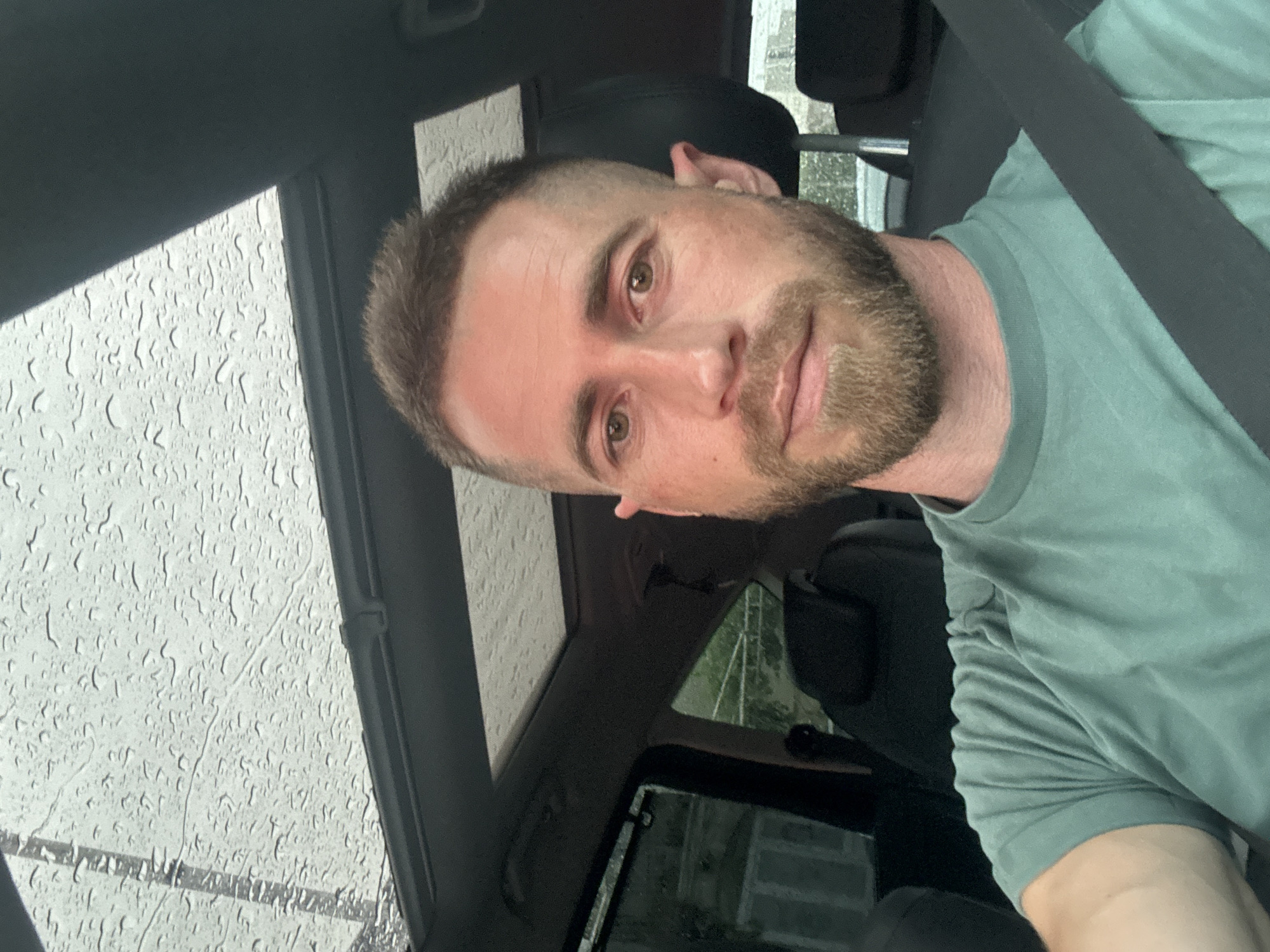 A man with a short, light brown haircut and beard is sitting in the driver's seat of a car. He is wearing a light green t-shirt and a seatbelt. It appears to be raining, as there are water droplets on the car's sunroof window. The car is likely located in Tivat, Montenegro based on the surrounding architecture and landscape. More specific location details are unavailable.