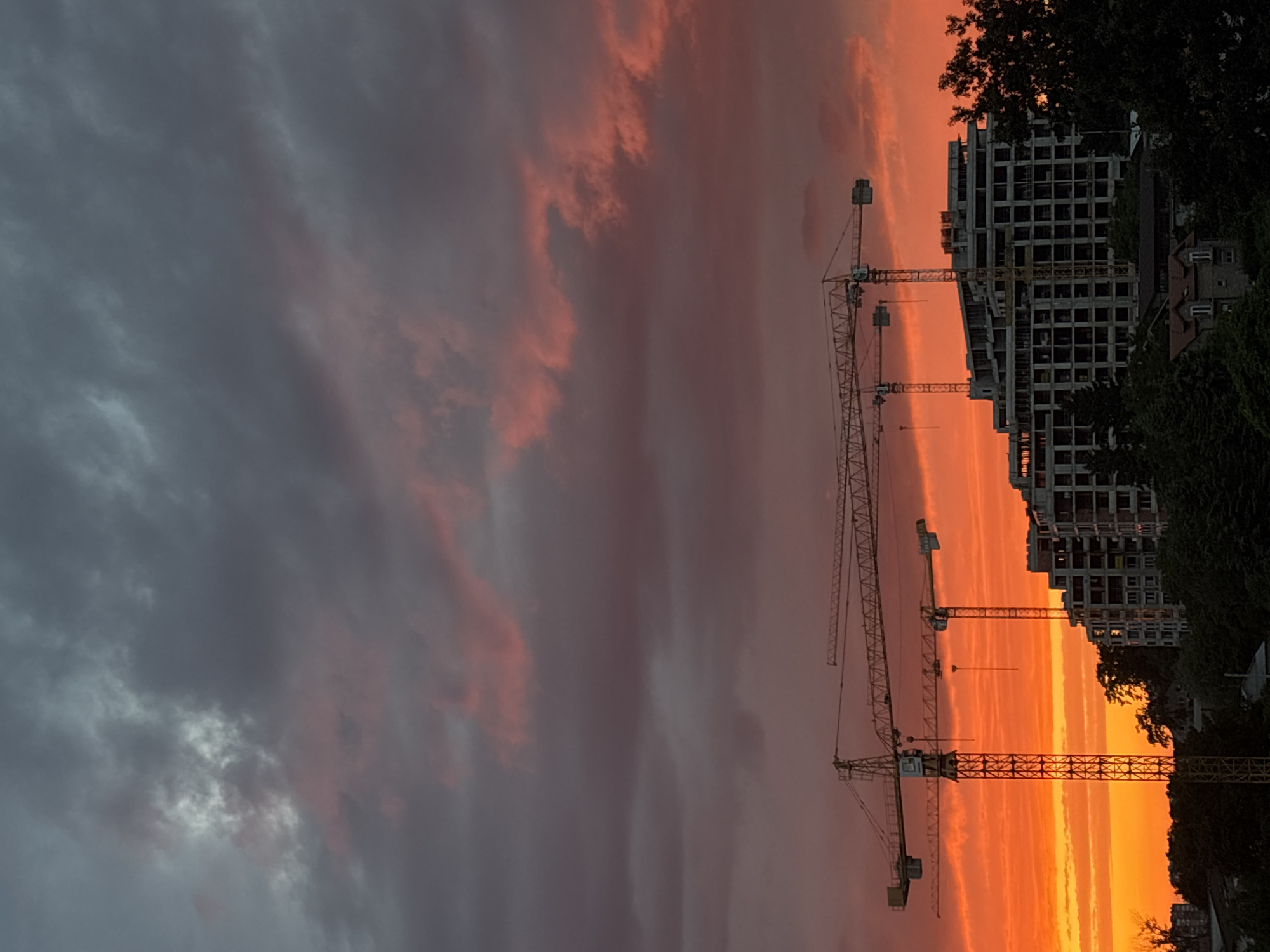 A vibrant sunset paints the sky with fiery oranges, pinks, and deep grays over a construction site in Zvezdara Urban Municipality, Serbia. In the foreground, two towering yellow construction cranes dominate the scene, their skeletal structures silhouetted against the dramatic sky. Behind them, a multi-story building under construction rises, its concrete frame stark and unfinished. Trees and smaller buildings form a dark silhouette at the base of the construction site. The image captures a moment of urban development against a backdrop of natural beauty.