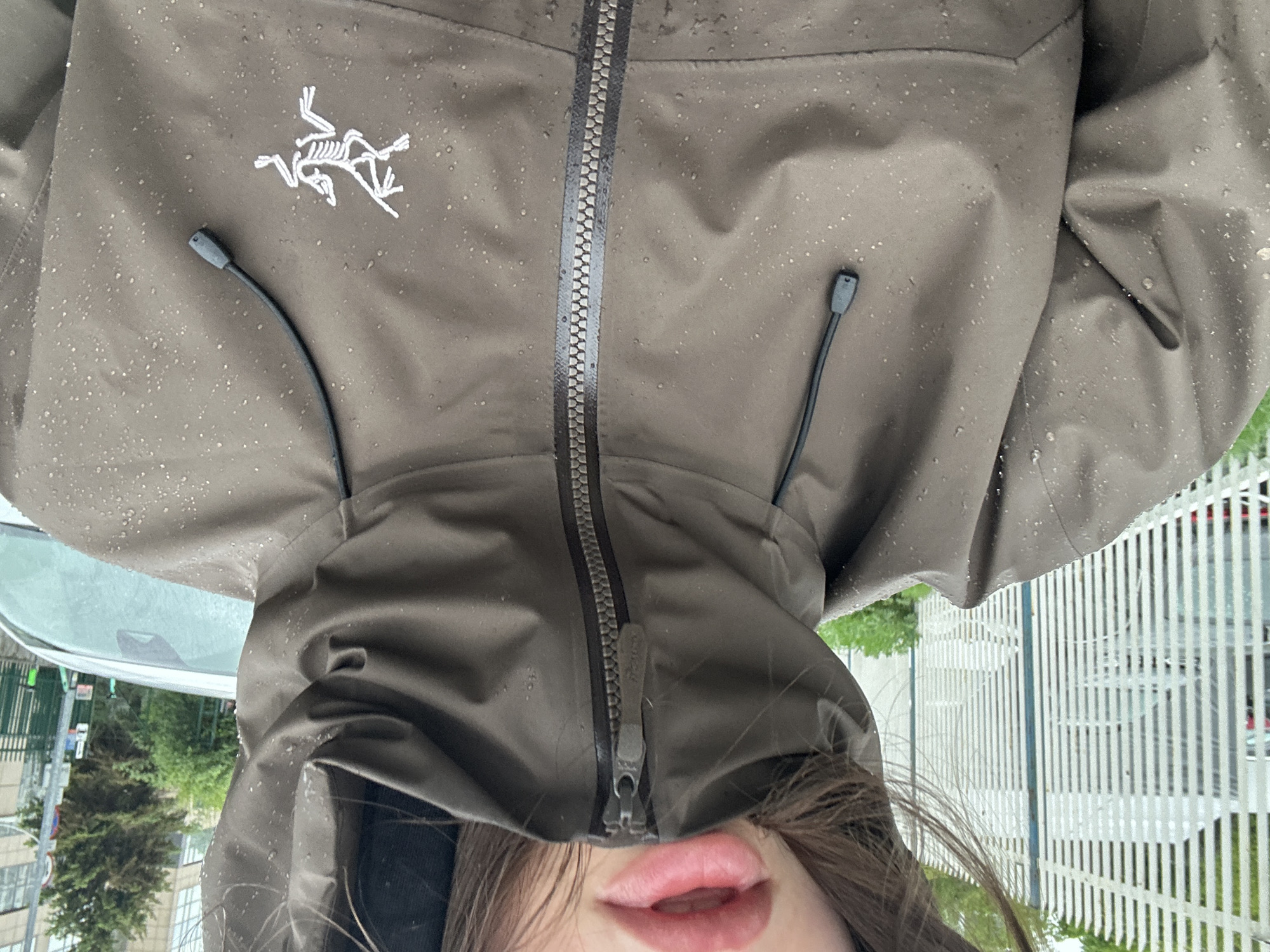 A person wearing a green Arc'teryx hooded jacket stands in front of a white picket fence. The jacket is zipped to the chin, and water droplets are visible on the fabric. The person's lips and hair are visible beneath the hood. A car, buildings, and trees are visible in the background. The location appears to be an urban area, possibly in the city of Unknown, Unknown.