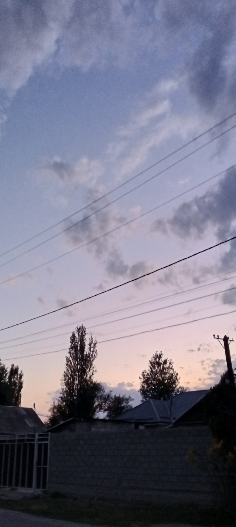 A low-angle, medium shot captures a dusky sky over the town of Sokuluk, Kyrgyzstan. The sky is filled with a blend of pale blue and streaks of gray clouds, with faint pink hues near the horizon. Several parallel power lines cut horizontally across the frame, obscuring parts of the sky. Below the power lines, the silhouettes of trees are visible against the twilight sky. In the foreground, a low, gray brick wall runs horizontally, with a dark metal gate on the left side. The roofs of houses are visible behind the wall, along with a utility pole on the right. The overall scene depicts a quiet, late evening in a residential area.