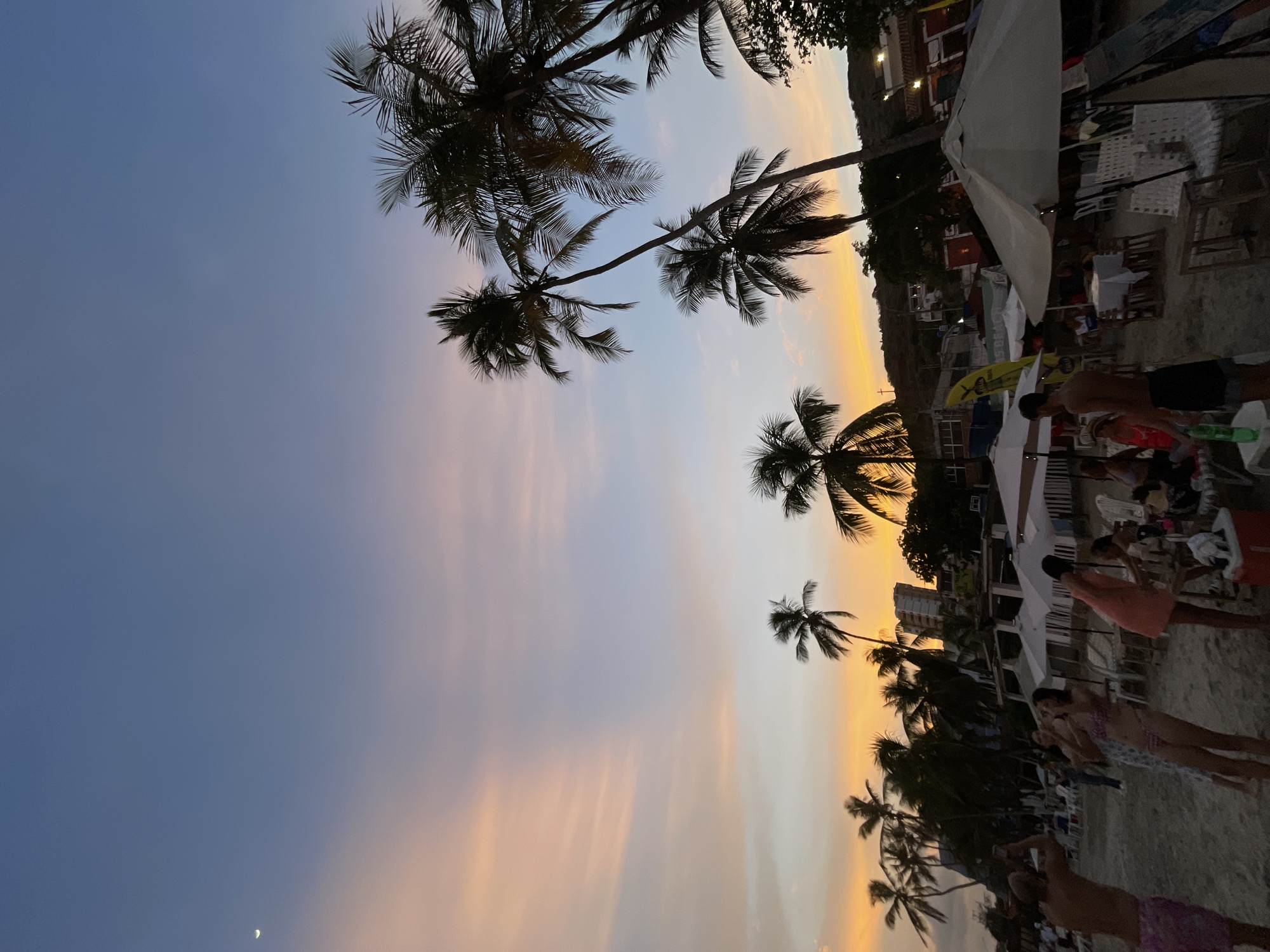 This image captures a sunset scene on a beach, likely in Pampatar, Venezuela. The sky is painted with hues of orange, pink, and blue, with a sliver of moon visible in the upper left corner. Palm trees are silhouetted against the colorful sky. In the foreground, several people are gathered on the sandy beach, some are taking pictures, while others are sitting at tables under umbrellas. There are beach chairs and tables set up, suggesting a casual dining or relaxation area. In the background, buildings and hills are visible, indicating a coastal town setting. The overall atmosphere is serene and leisurely, typical of a tropical beach at dusk.