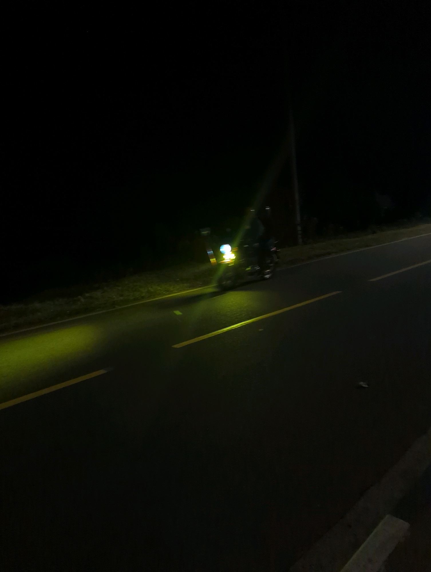 An underexposed, low-light photograph captures a motorcycle traveling on a paved road at night. The motorcycle's headlights cast a bright yellow glow on the road surface, illuminating a section of the asphalt and the yellow lane markings. The motorcycle itself is partially visible, with a rider on board, but is mostly obscured by the darkness and the intense headlight beams. The background is predominantly black, with a faint indication of vegetation along the side of the road and a vertical utility pole or street light. The scene appears to be a rural or semi-rural road within Musanze District, Rwanda, given the context provided. The image suggests motion and the isolated nature of nighttime travel on this road.