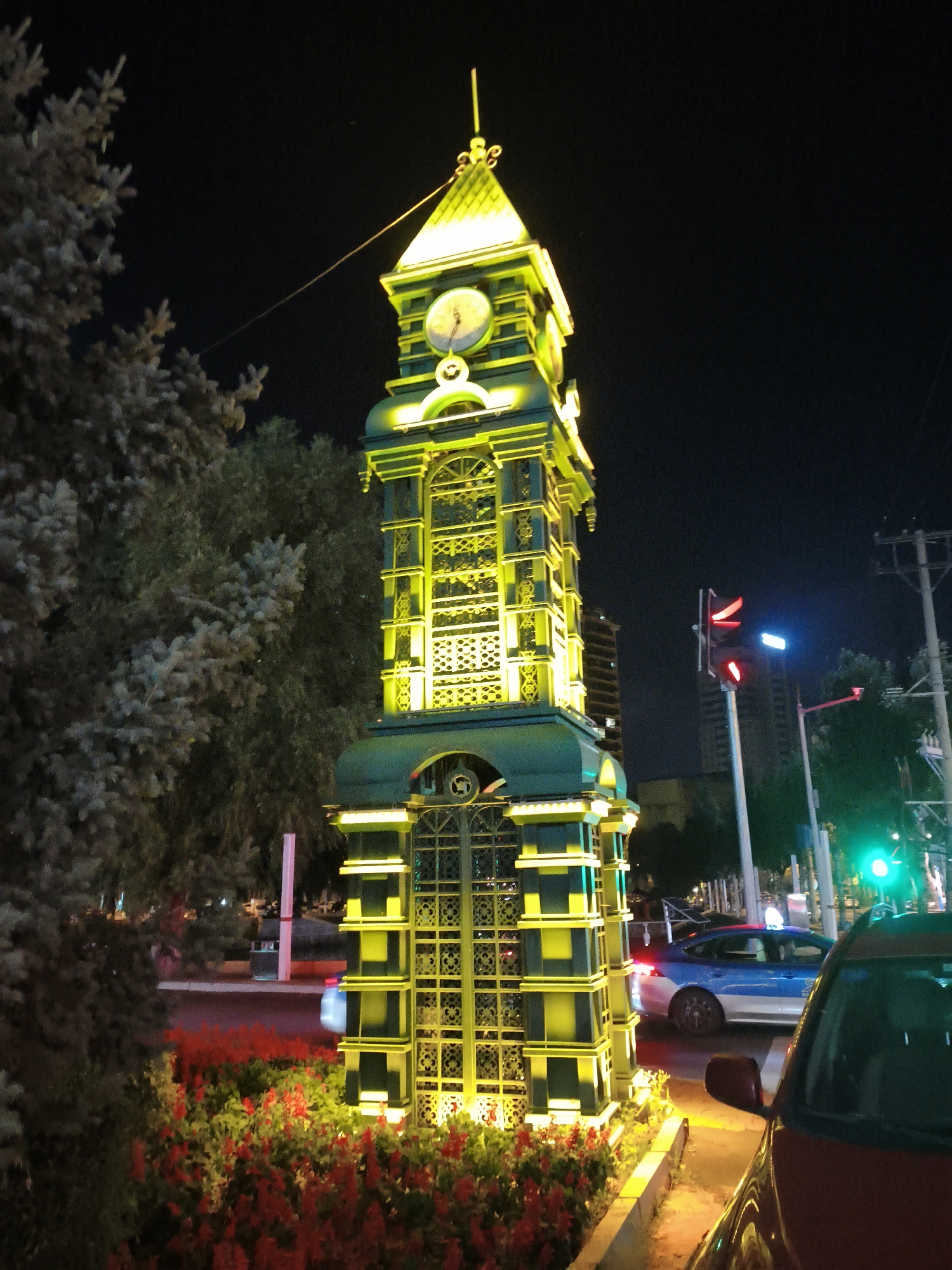 The image shows a night scene of a decorative clock tower in the city of Xi'an, China. The clock tower is tall and narrow, with multiple levels adorned with intricate patterns and illuminated by yellow lights. A clock face is visible near the top of the tower. The tower is situated in an urban environment, with trees and flowering plants in the foreground and a street with traffic in the background. A blue and white car is visible on the street, and a red car is parked in the foreground on the right. Traffic lights are also visible, one showing red and another showing green. The sky is dark, indicating it is nighttime. The overall impression is of a well-lit, vibrant city scene at night.