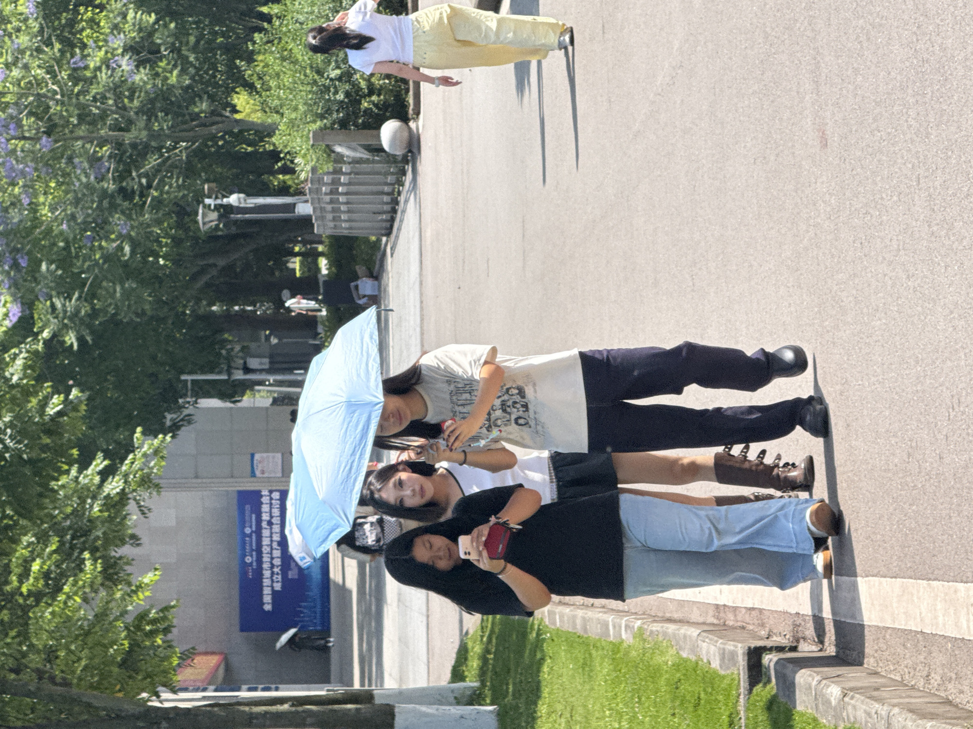 Three girls pose for a photo. One in the front, wearing a black shirt and light blue jeans, holds a phone. The second girl, in a white tank top and black skirt, smiles. The third girl, in a tan t-shirt and dark pants, holds a bottle. A light blue umbrella is held over them. A woman in a white shirt and yellow pants walks away in the background. The scene appears to be in an urban environment, possibly in Jiangjin, China, based on the Chinese characters on a blue sign nearby. The area includes trees, greenery, a concrete pathway, and street furniture.