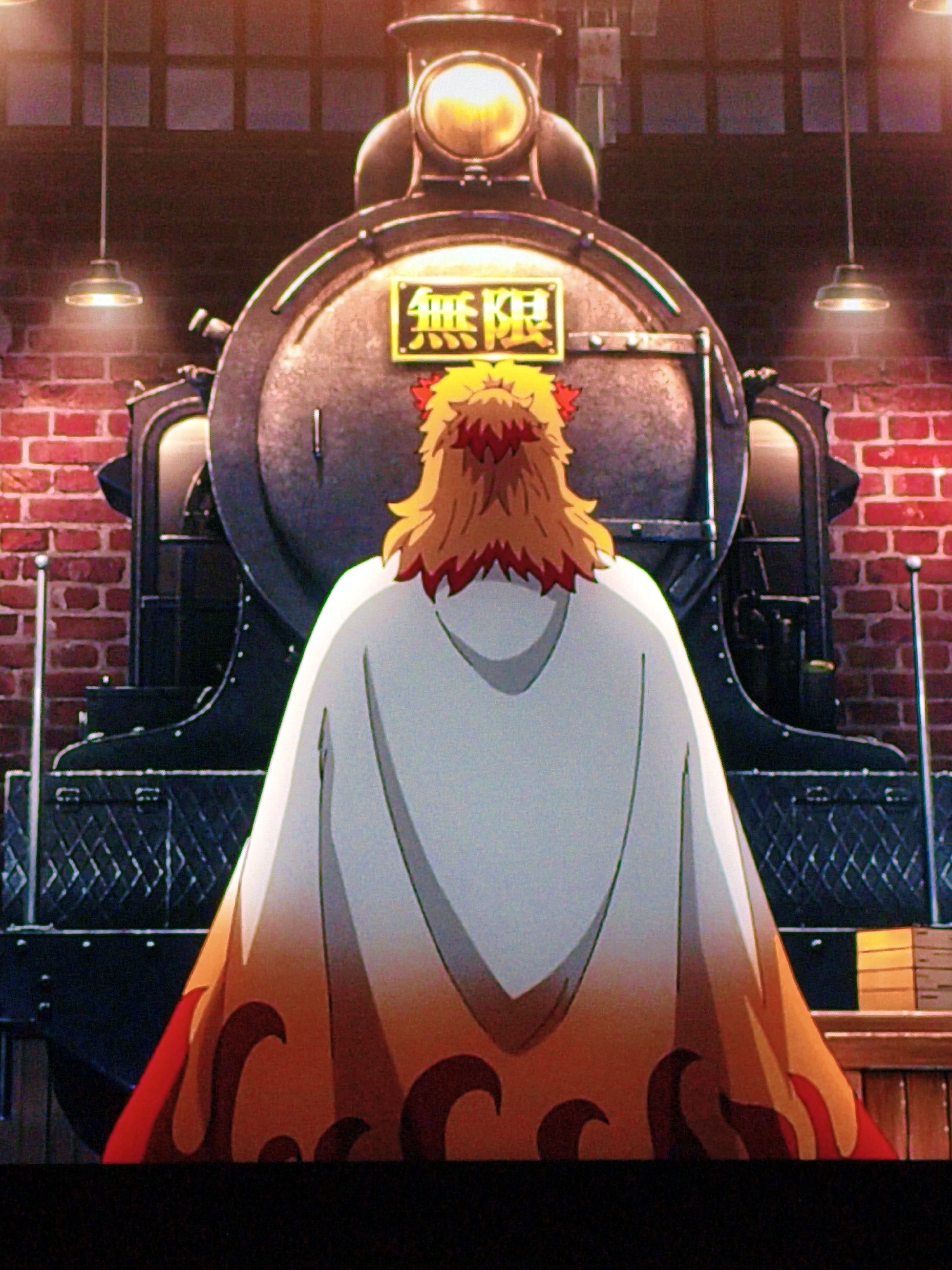 A low-angle, medium shot shows the rear view of an anime character with prominent orange and red flame-like designs on their cape, standing before the front of a large, dark steam locomotive. The character has spiky orange hair and a red hair accessory. The locomotive has a large, illuminated headlight at the top, and a yellow rectangular sign with Japanese characters (presumably "Mugen" or "Infinity") is affixed to its boiler. The background depicts a brick wall with visible mortar lines, and industrial-style hanging lamps with exposed bulbs. Wooden crates are stacked to the right of the frame. The scene appears to be a still from an anime or animation, given the artistic rendering. There is no indication that this scene is located in Vladivostok, Russia, or anywhere else specific outside of the fictional context.