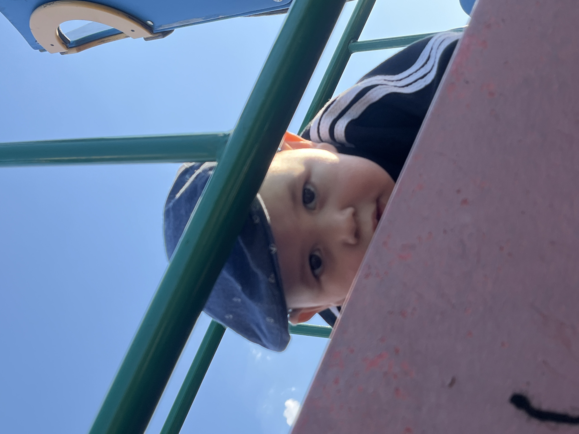 A low-angle shot shows a baby wearing a blue cap and a black jacket with white stripes peering down from a playground structure. The structure is made of green metal bars and a pink surface. The sky is visible in the background. The location in the city of Фоки, Russia is undetermined.