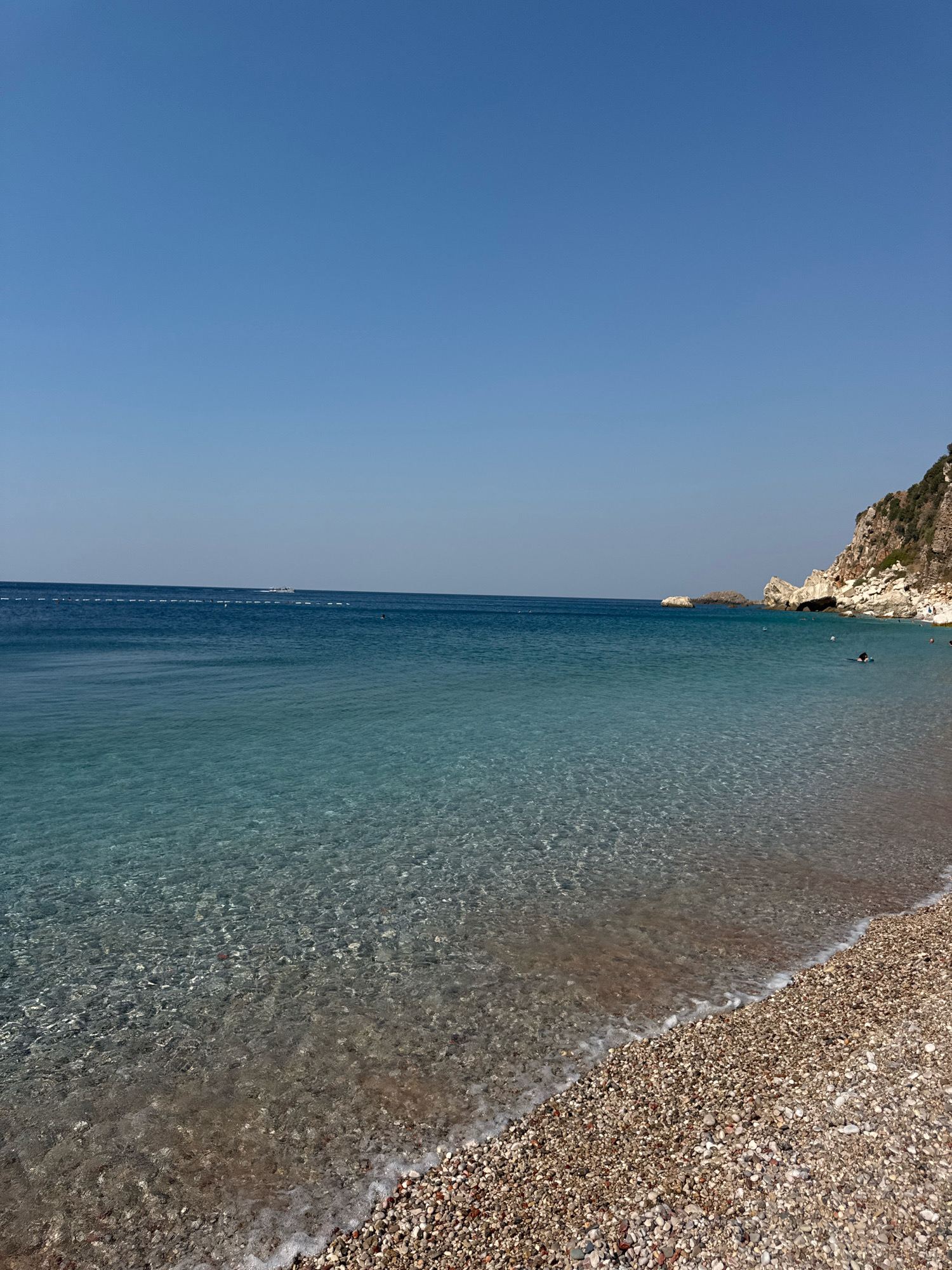 The image depicts a scenic view of a pebble beach and the clear, turquoise waters of the Adriatic Sea in Mirište, Montenegro. The sky is a clear, bright blue, indicating a sunny day. The foreground is dominated by the pebble beach, with small, rounded stones of various colors. Gentle waves lap at the shore, creating a thin line of white foam.
The water is exceptionally clear, allowing the seabed to be visible. The water transitions from a lighter, translucent hue near the shore to a deeper blue further out. In the distance, there is a faint line of buoys, possibly marking a swimming area. Further still, a small boat can be seen on the horizon.
To the right of the frame, a rugged, rocky cliff face rises from the sea. The cliff is covered in sparse vegetation, and its rocky texture is clearly visible. Several large rocks and formations are scattered in the water near the cliff.
There are a few people visible in the water, swimming and enjoying the sea. One person is clearly visible swimming near the rocky outcrop. The overall impression is one of a tranquil and beautiful coastal landscape.