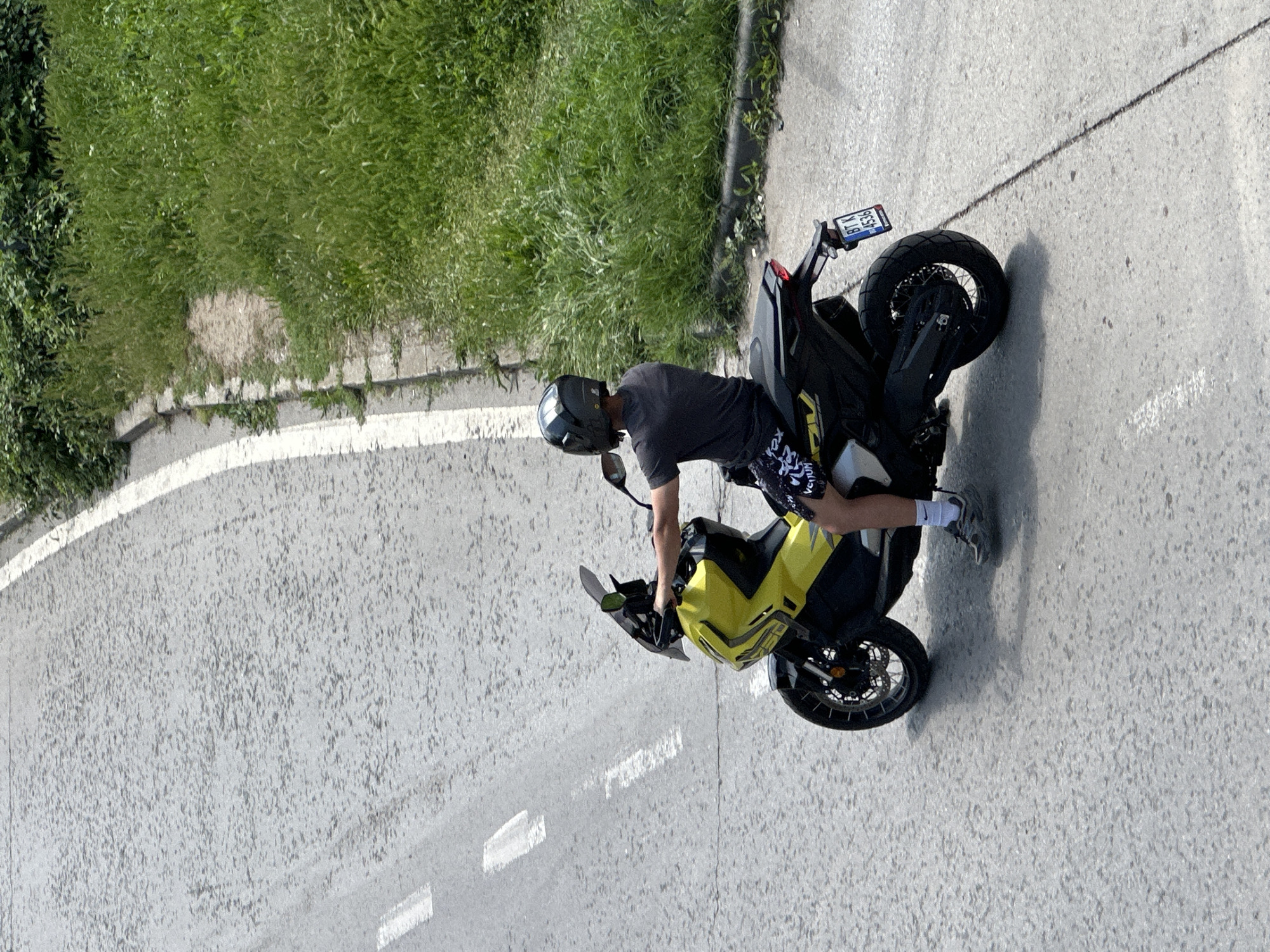 The image depicts a person on a yellow and black motorcycle parked on a paved road during what appears to be a bright, sunny day in Veliko Tarnovo, Bulgaria. The perspective is from an elevated position, looking down at the scene.
The central subject is a male individual, wearing a dark grey t-shirt, black shorts with a white and yellow pattern (possibly featuring "Venum" text), white socks, and grey athletic shoes. He is also wearing a dark grey or black full-face motorcycle helmet. The person is seated on the motorcycle, with his right leg extended towards the ground, suggesting he is either stopped, balancing the bike, or in the process of mounting or dismounting.
The motorcycle itself is a distinctive yellow and black model, possibly an adventure or supermoto style, featuring spoked wheels. Its rear fender holds a clearly visible license plate, which displays "BT K 4536". The "BT" prefix identifies the registration as being from Veliko Tarnovo, Bulgaria, aligning with the provided location context.
The setting is an outdoor road with a slight curve. The road surface is grey pavement, likely asphalt, marked with white painted lines including a solid line along the curb and dashed lines further into the lane. To the left of the road, a concrete curb separates the pavement from a vibrant green grassy bank, indicating a natural roadside environment. Bright sunlight casts clear shadows, confirming it is daytime with good weather conditions.