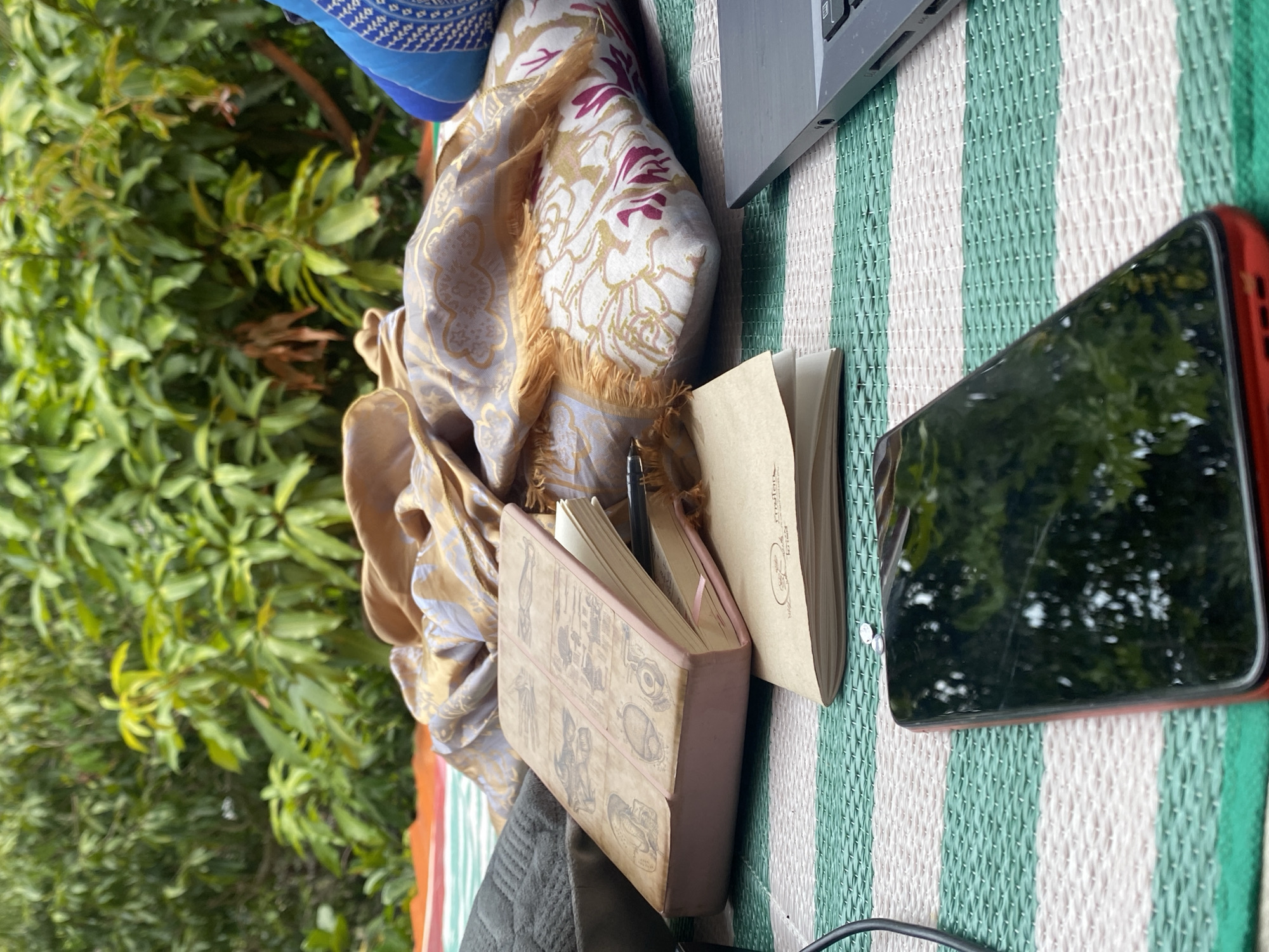 The image shows a picnic-like arrangement outdoors, possibly in Baan Kai, Thailand, based on the surrounding vegetation which includes lush green foliage. A striped mat in alternating green and white is laid out, upon which several items are placed. There is a laptop with its keyboard partly visible, alongside a red-cased smartphone reflecting the surrounding greenery. Two open notebooks lie on the mat; one with plain pages and a tan cover, the other with anatomical drawings on its pink cover. A partially visible blue patterned object and a draped cloth with gold and white patterns are also present.