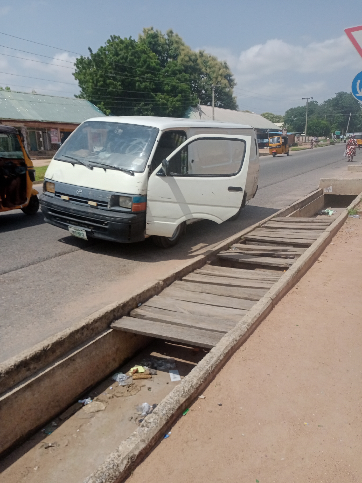 This is a street-level, medium shot photograph taken in Jalingo, Nigeria, on a cloudy day. In the foreground, a concrete drainage channel runs along the side of the road. The channel is partially covered by wooden planks, some of which are broken or missing, creating a makeshift bridge. Debris, including plastic bags and other litter, is visible within the drainage channel.
A white minibus, with its driver's side door open, is parked on the asphalt road, partially blocking the view of the road ahead. The minibus has a license plate that appears to read "MNT 9853". To the left of the minibus, the front of a yellow motorized tricycle is visible.
Further down the road, more vehicles and pedestrians are present. A yellow motorized tricycle is in motion. Two women, one wearing a patterned yellow and white dress and the other in a blue and white checkered dress, are walking on the sidewalk on the right side of the road. A white traffic sign with a red border and a blue circular sign with a white symbol are visible on a pole on the right side of the image.
The background shows a row of buildings with metal roofs, lush green trees, and utility poles with overhead wires. The sky is partly cloudy. The overall scene depicts a busy street in an urban environment with signs of disrepair in the infrastructure, such as the damaged drainage cover.