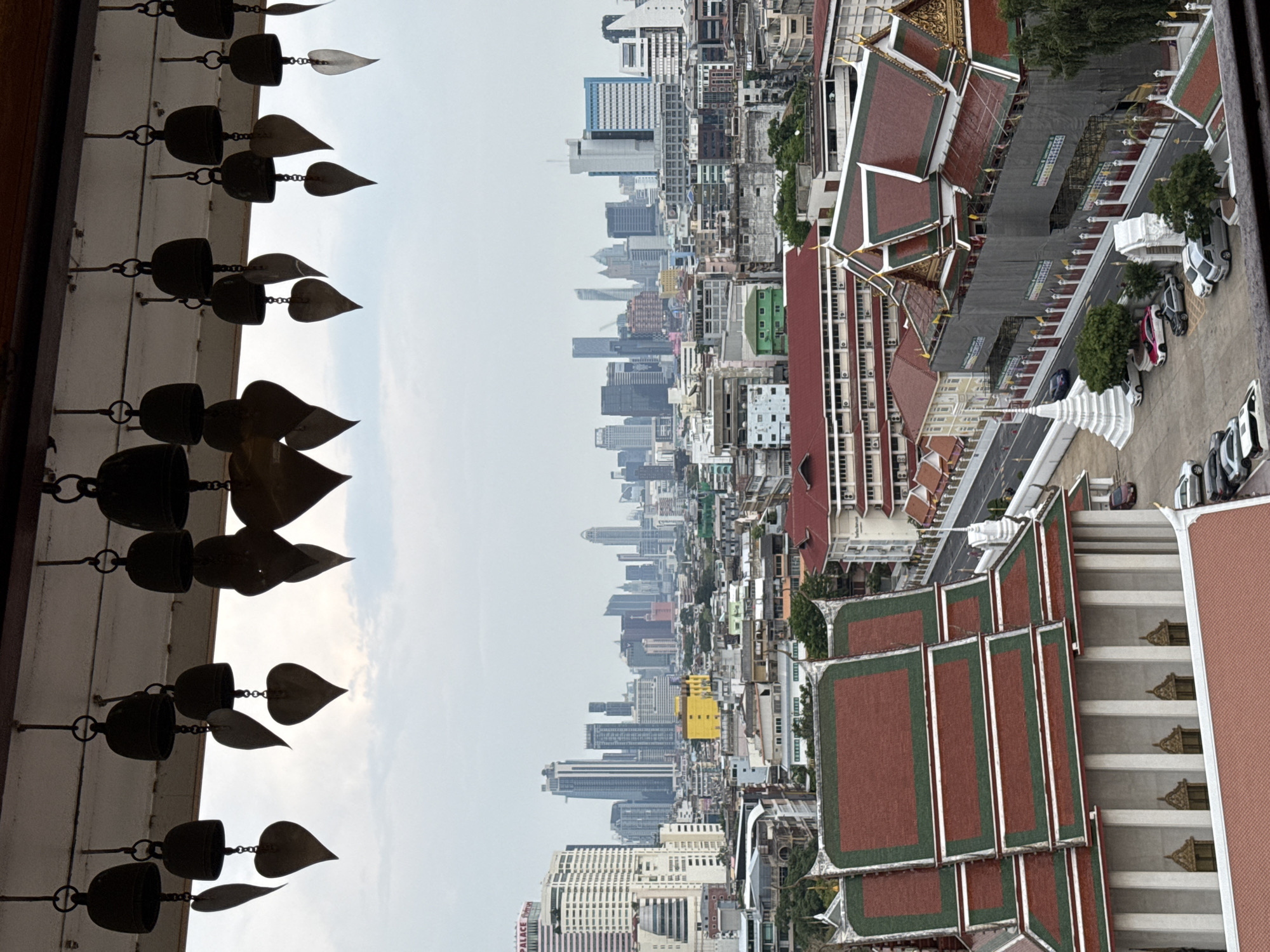The image depicts a partial view of Bangkok, Thailand, with a focus on elements of a temple and the urban cityscape. On the left, rows of hanging bells and metallic leaf ornaments frame the edge. The center of the image shows a cityscape with varying building heights and architectural styles, typical of urban Bangkok. In the foreground, a temple rooftop with red tiles and ornate decorations dominates the right side. A staircase is visible, suggesting an elevated viewpoint. A street with parked cars and surrounding greenery are also visible. The sky is overcast.