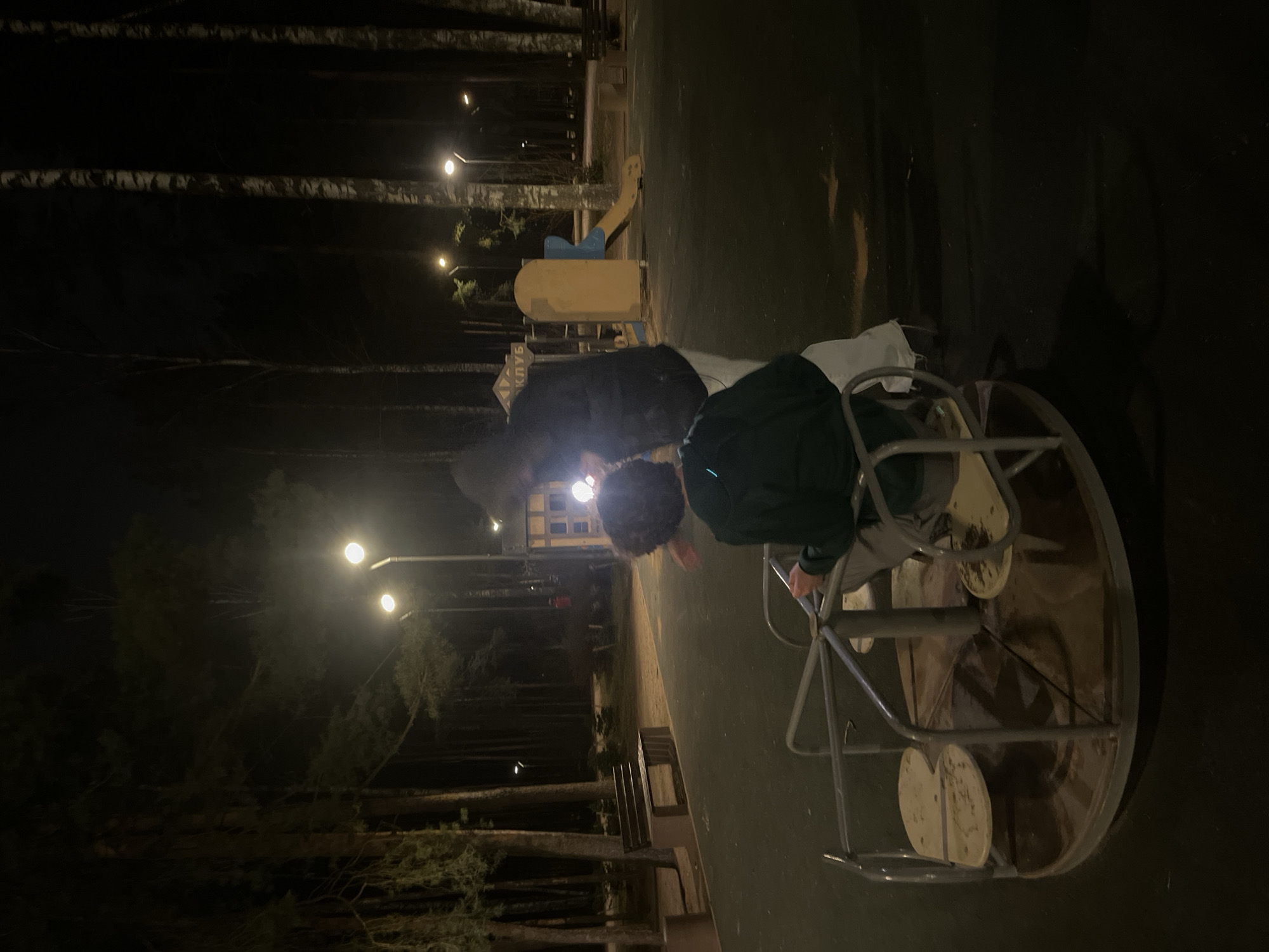 A playground scene at night in what is likely Saint Petersburg, Russia. Two people are interacting with a merry-go-round. One person is sitting on the rotating part of the merry-go-round, and the other person is pushing it. The scene is lit by street lamps. A playground set is visible in the background. Trees line the edges of the playground. There are benches in the foreground.