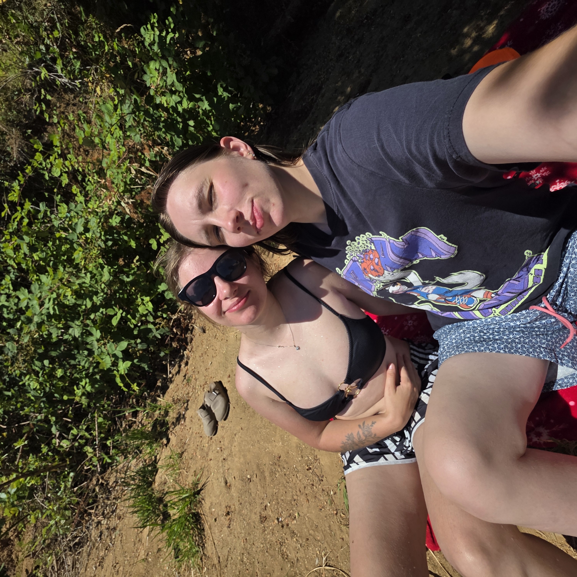In Cherry Grove, Oregon, two individuals are captured in a selfie taken outdoors. They are positioned on the ground, with lush green foliage in the background and a sandy, earthy terrain surrounding them. The person on the left is wearing a black bikini top and patterned bottoms, with sunglasses resting on her head. The person on the right is wearing a dark t-shirt with a graphic print. Both appear relaxed and are enjoying the natural environment. A pair of casual shoes are visible in the background, suggesting they may have been walking or hiking prior to taking this photo. The lighting indicates it is daytime, with sunlight filtering through the trees, creating dappled shadows.