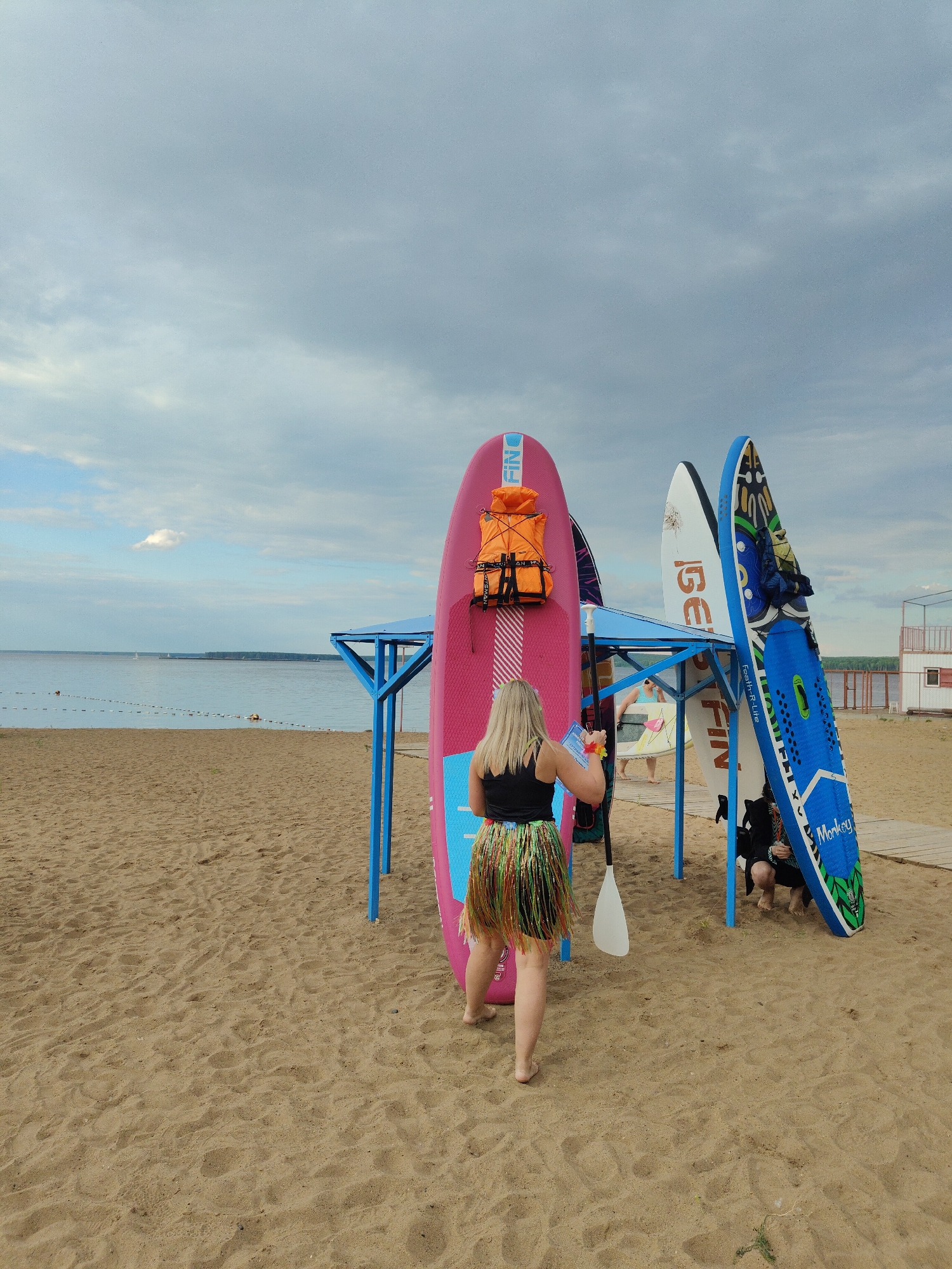 A group of people are on a sandy beach near a body of water. In the foreground, a woman with blonde hair wears a black tank top and a colorful, fringed skirt. She is holding a piece of paper and a lei in her hands, facing away from the camera. A white paddle is leaning against a pink stand-up paddleboard (SUP) next to her. This SUP has an orange life vest attached to it. Behind the woman, several other SUPs are propped up against a blue metal structure, which appears to be a covered shelter. One of these SUPs is blue with green and black patterns and text that reads "Monkey". Another SUP is white with orange text that reads "GEFIN". Further in the background, a few people are visible near the water and a white building with a red roof is situated on the beach. The water is calm and appears to be a lake or a large bay, with a few boats visible in the distance. The sky is overcast with some patches of blue, suggesting a cloudy day. The scene appears to be a recreational area, possibly a rental spot for water sports equipment.